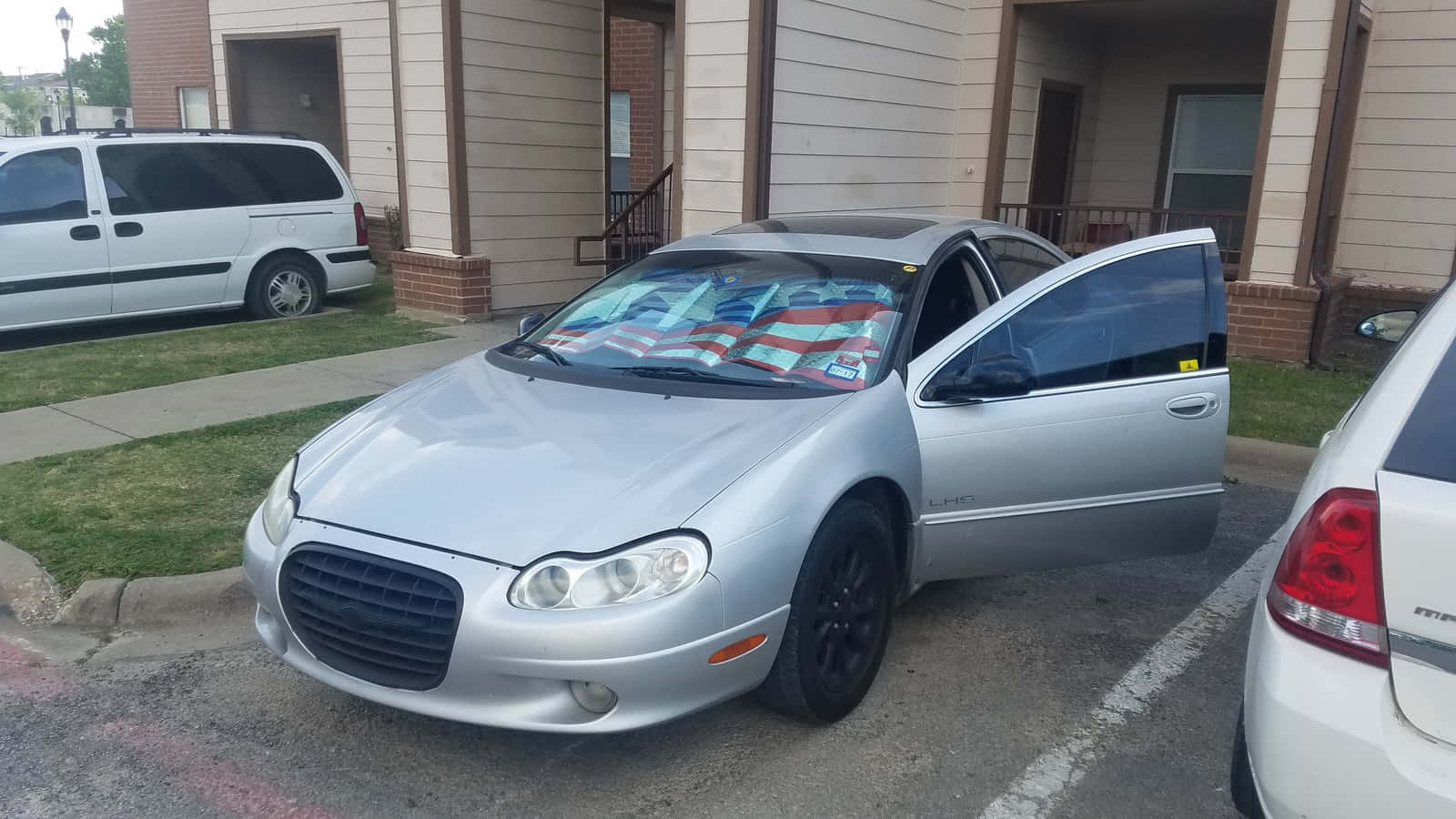 Chrysler Lhs 桌布 壁紙 — 一輛銀色克萊斯勒Sebring，駕駛座車門打開，美國國旗披在擋風玻璃上，停在住宅區。