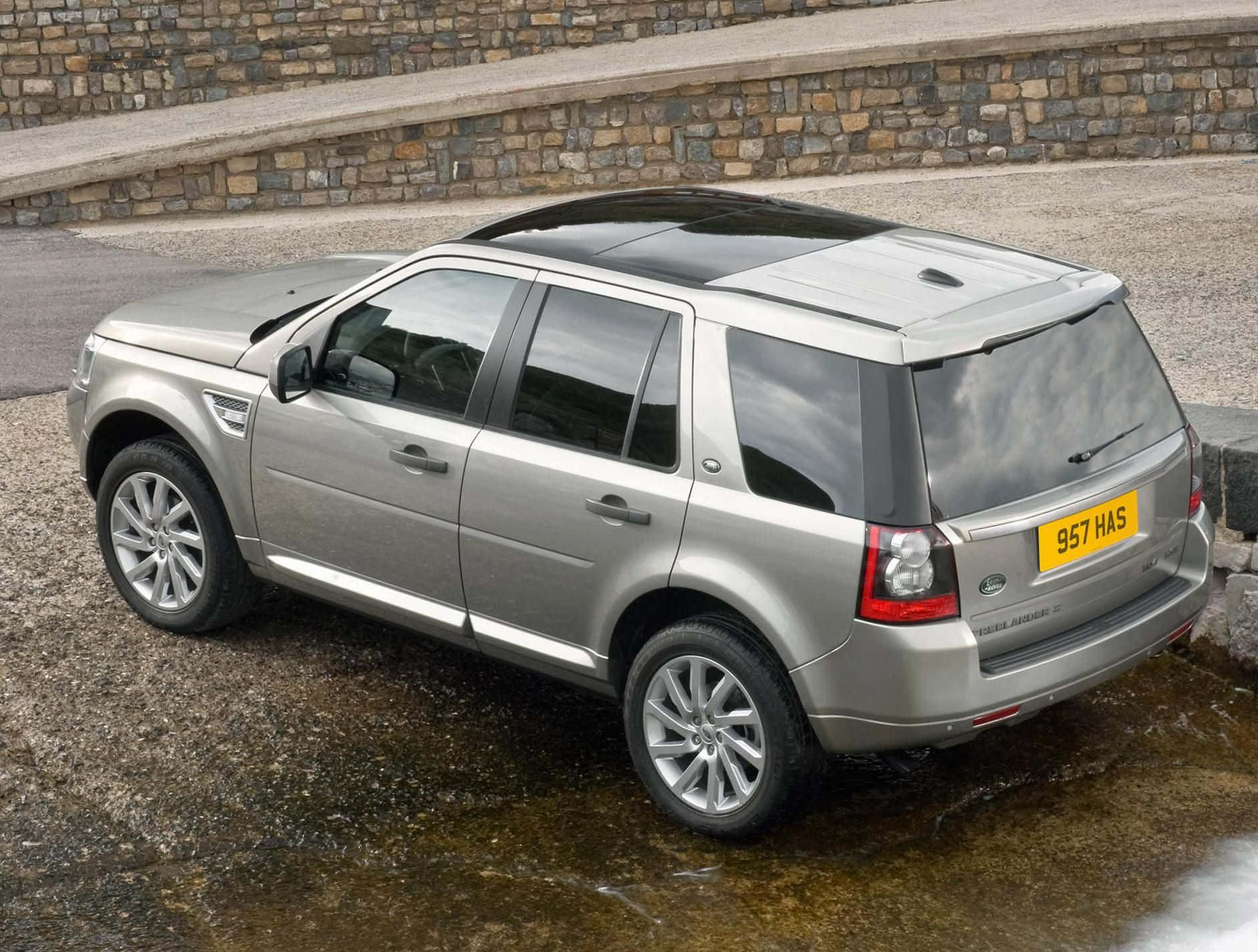 Land Rover Freelander 桌布 壁紙 — 一輛銀色 Land Rover Freelander 2 停在濕碎石上，旁邊有石牆，輪胎周圍積水。