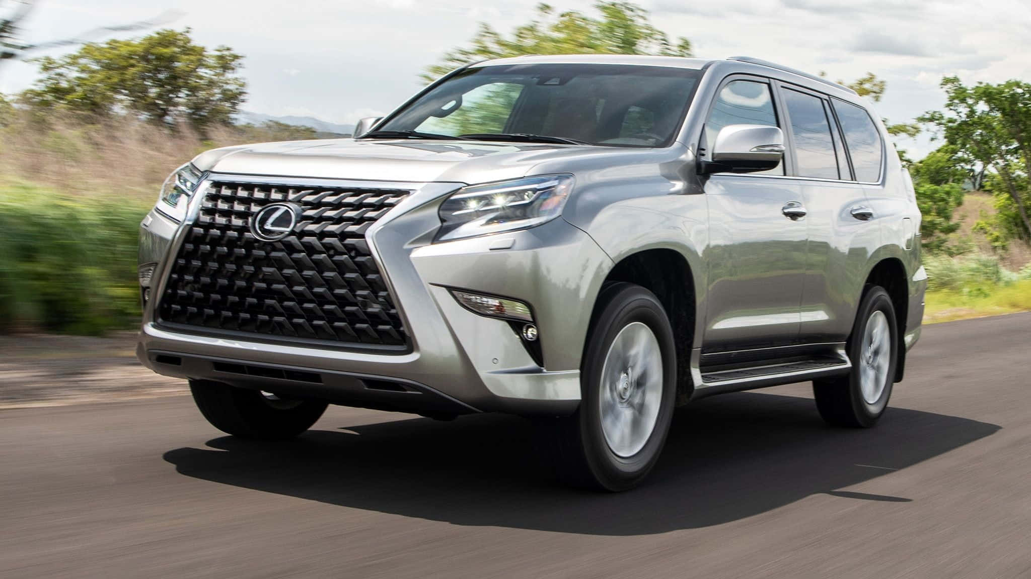 Lexus Gx 460 桌布 壁紙 — 一輛銀色Lexus GX SUV在陽光普照的道路上行駛，背景是模糊的綠樹和遠山。