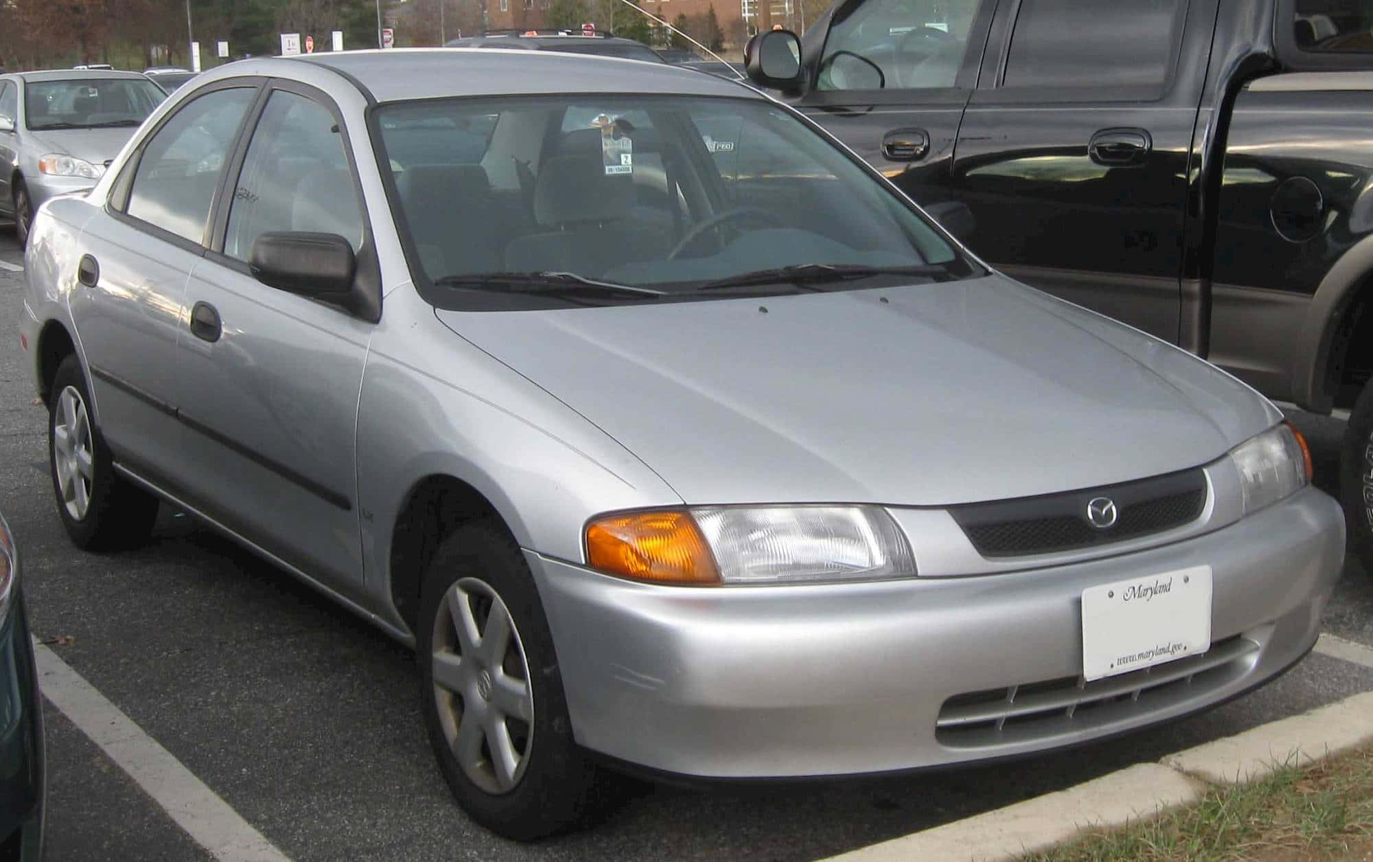 Mazda Protege 桌布 壁紙 — 一輛銀色馬自達轎車停放在戶外停車場，附近有其他車輛。