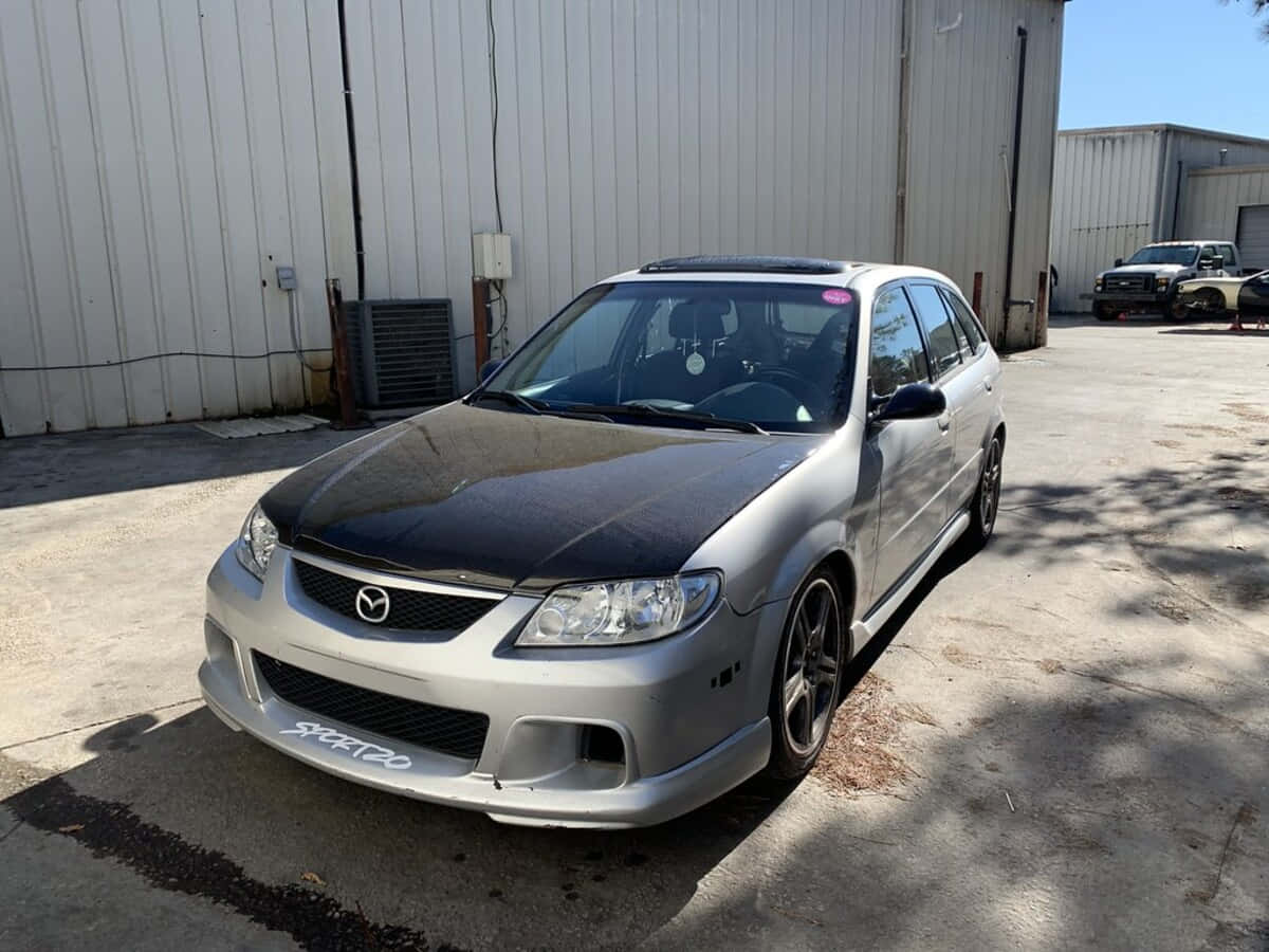 Mazda Protege 桌布 壁紙 — 一輛銀色Mazda 3，黑色引擎蓋和客製化輪圈，停在金屬倉庫前，陽光普照。