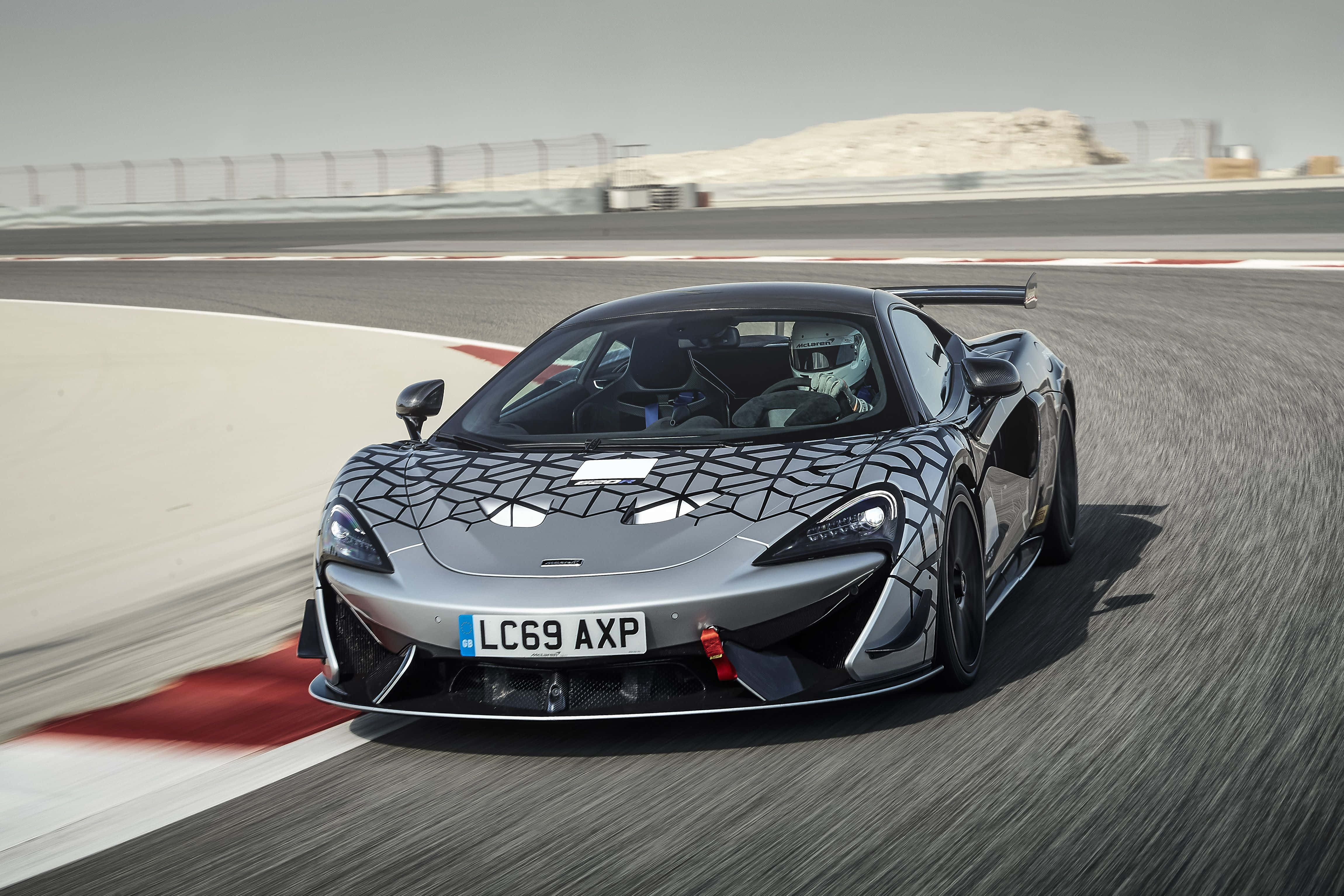 Mclaren 620r 桌布 壁紙 — 一輛銀色McLaren跑車帶有黑色幾何迷彩，在明亮的日光下於賽道彎道疾馳。
