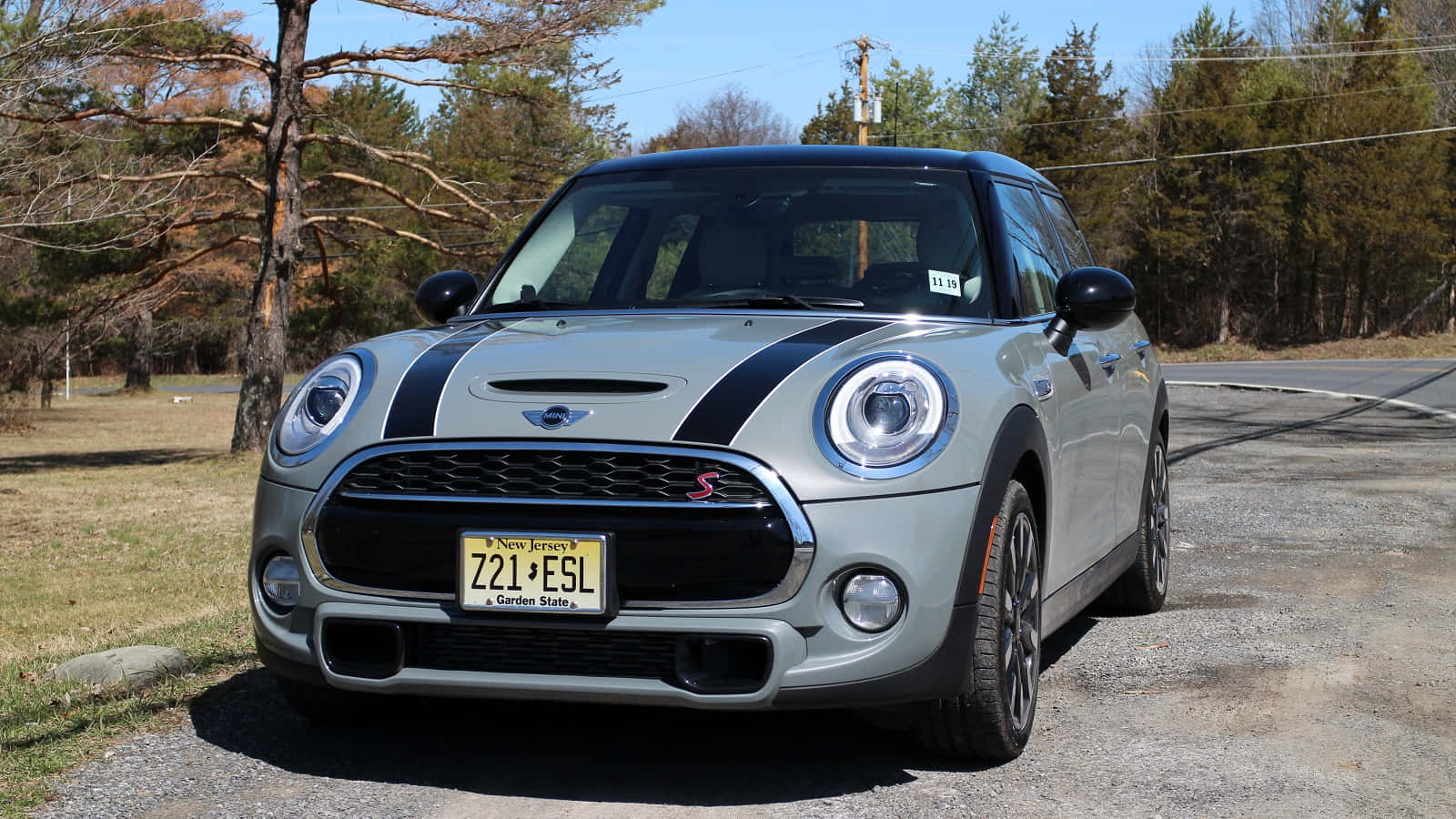 Mini Cooper Hardtop 2 Door 桌布 壁紙 — 一輛銀色MINI Cooper S帶有黑色賽車條紋，停在碎石路上，旁邊是樹木成蔭的道路，天空清澈湛藍。