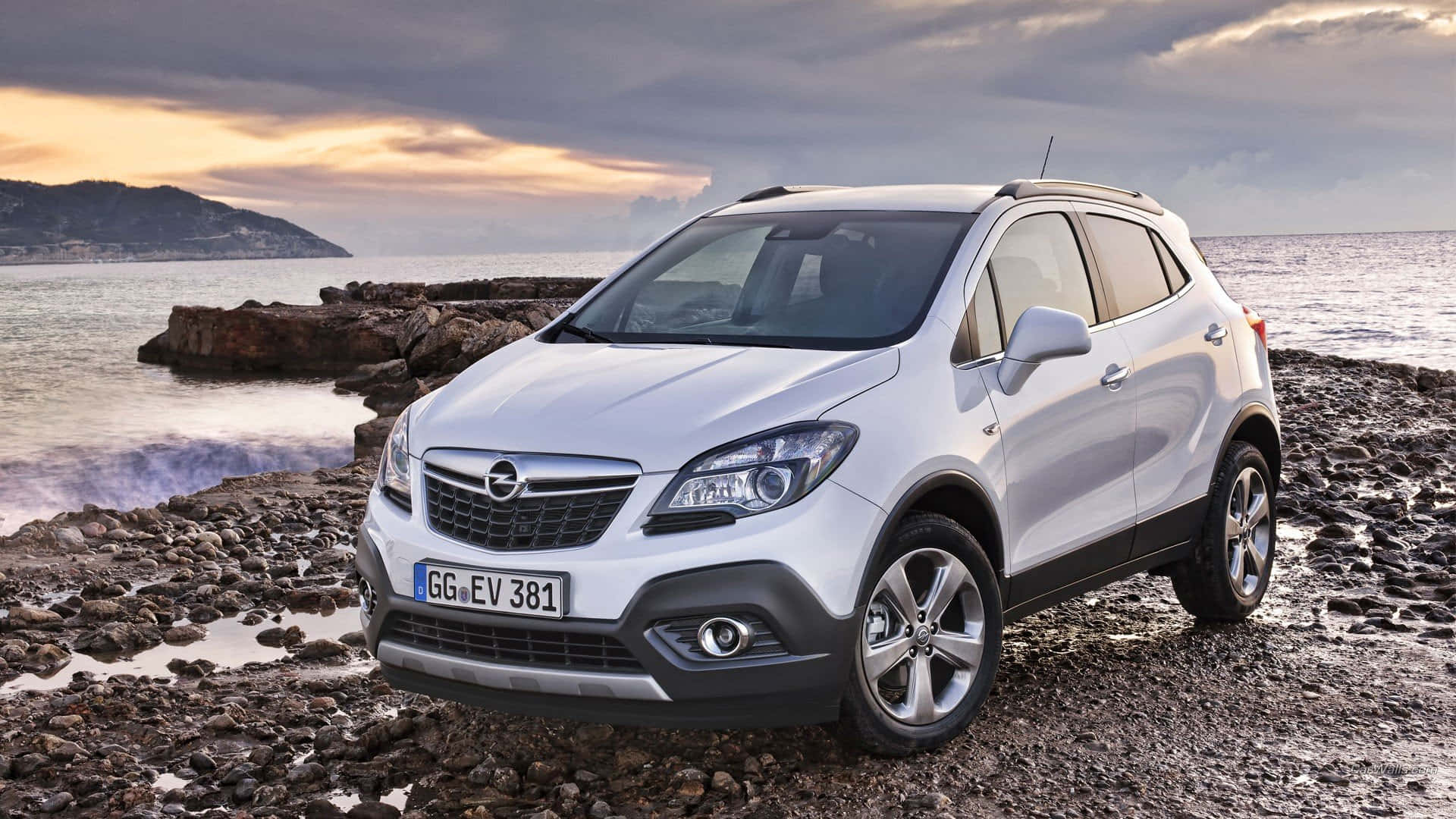 Opel Mokka 桌布 壁紙 — 一輛銀色Opel Mokka SUV停泊在日落時分的岩石海岸，背景有戲劇性的雲層和海洋波浪。