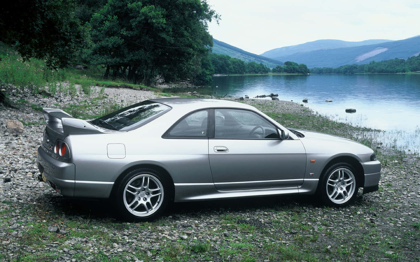 銀色跑車停泊在湖邊碎石上，後面有山。 — 日產Skyline GTR R33壁紙 壁紙