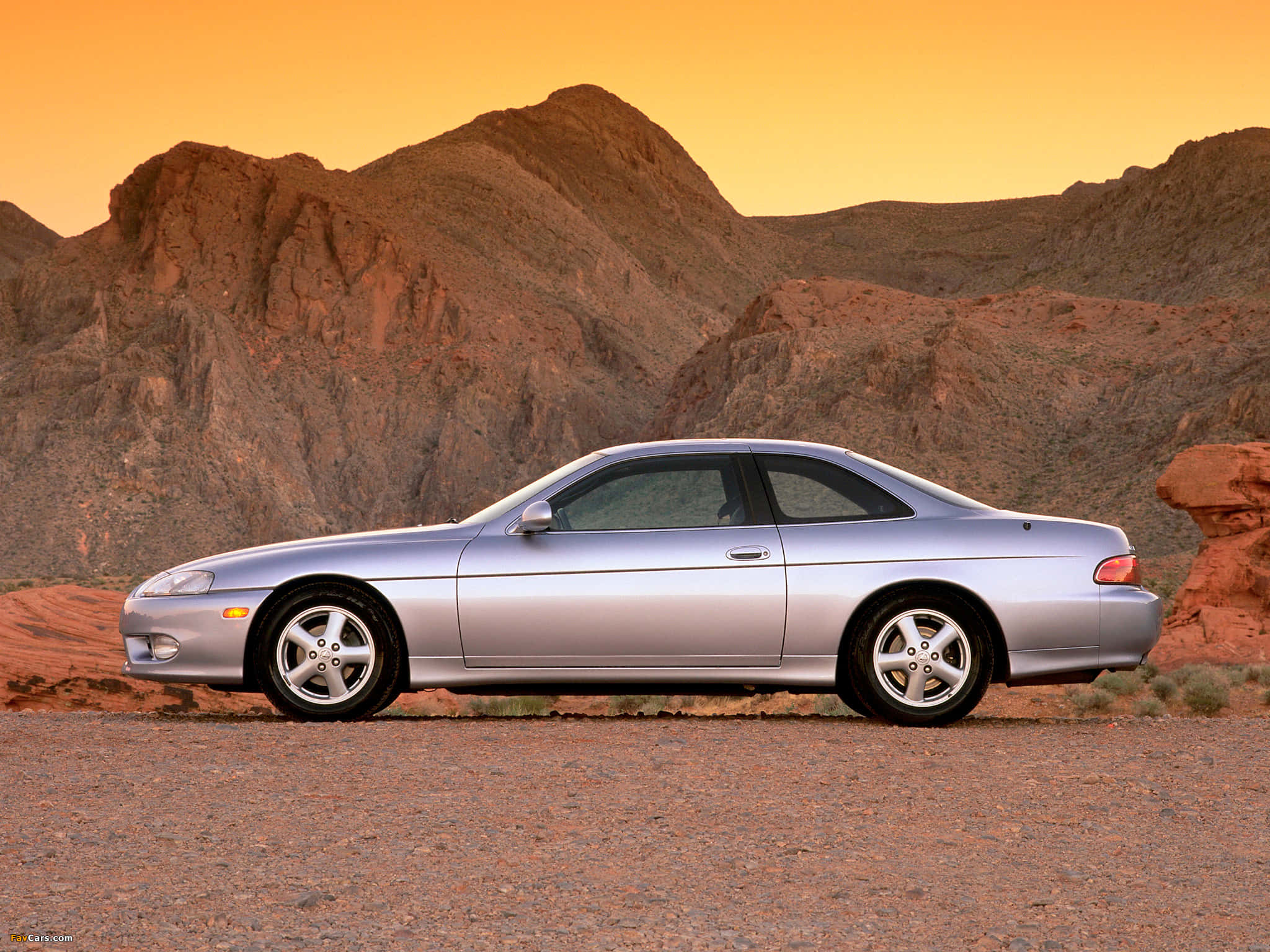 Lexus Sc 300 桌布 壁紙 — 一輛銀色跑車停在沙漠公路上，背景是紅色岩石地貌，天空呈現溫暖的橙色日落。