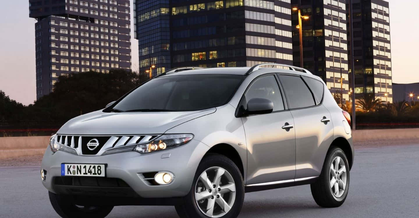 Nissan Murano 桌布 壁紙 — 一輛銀色日產Rogue SUV在黃昏時停放在現代玻璃摩天大樓前，窗戶亮著燈。