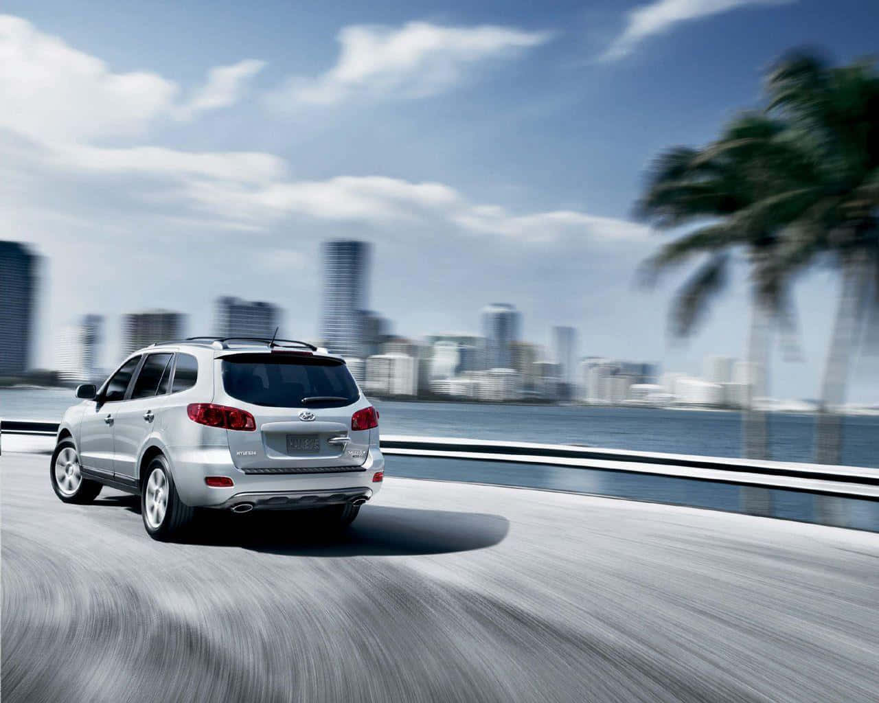Hyundai Santa Fe 桌布 壁紙 — 一輛銀色SUV沿著海岸公路飛馳，背景有棕櫚樹和模糊的城市天際線。