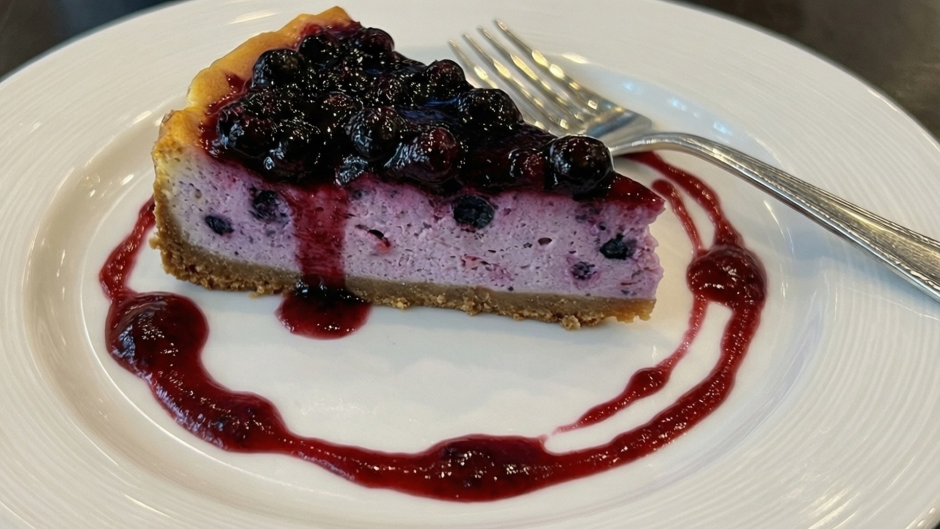 Blueberry Cheesecake Slice 壁紙 — 一片藍莓起司蛋糕，配上光澤的莓果和紅色醬汁，放在白色盤子上，附有叉子。