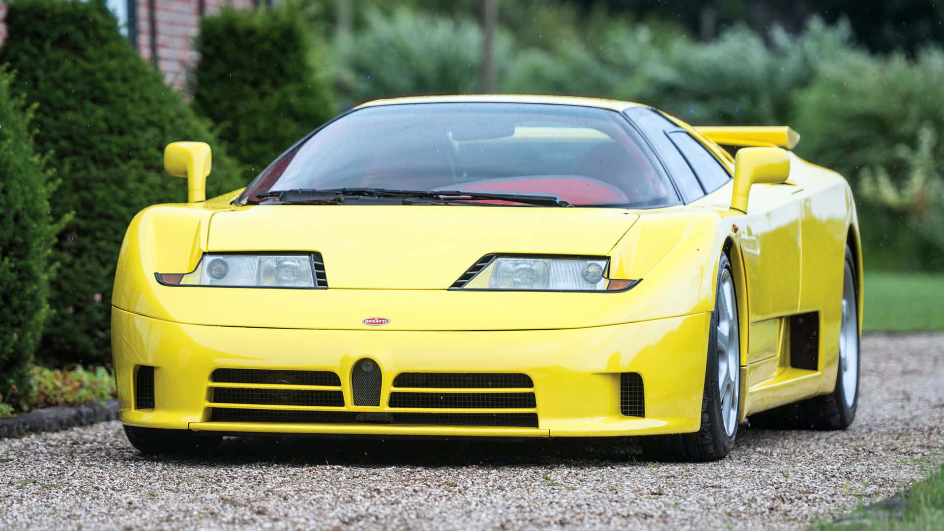 Bugatti Eb110 桌布 壁紙 — 一輛亮黃色的 Bugatti EB110 Super Sport 跑車停放在碎石車道上，周圍環繞著綠色灌木。