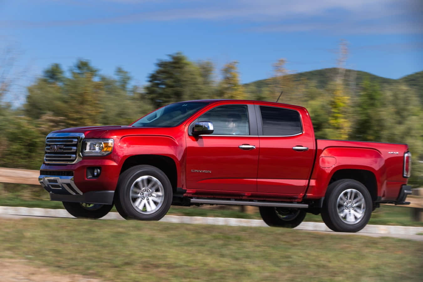 Gmc Canyon 桌布 壁紙 — 一輛充滿活力的紅色GMC Canyon皮卡車在路邊疾馳，背景是模糊的綠色樹木和山丘，天空清澈湛藍。