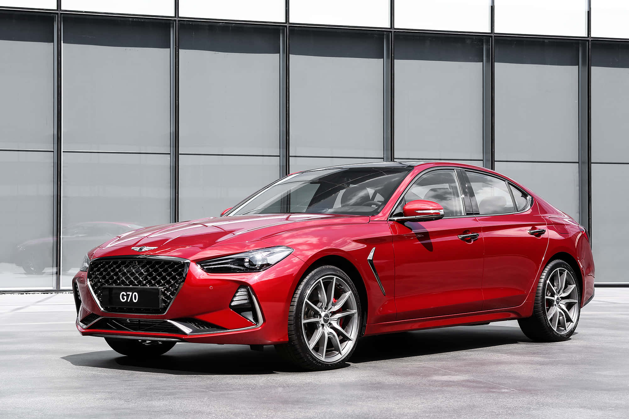 Genesis G70 桌布 壁紙 — 一輛亮紅色的現代 G70 轎車停在現代玻璃建築前，在明亮的日光下。