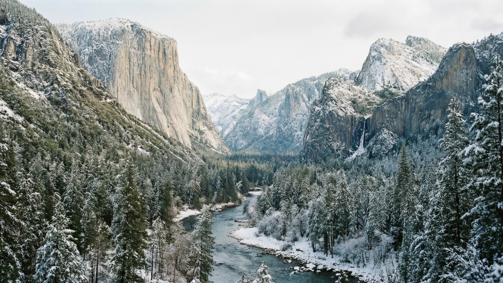Winter Yosemite Valley 壁紙 — 一條被雪覆蓋的河流蜿蜒穿過森林山谷，兩旁是高聳的花崗岩懸崖，天空陰沉。