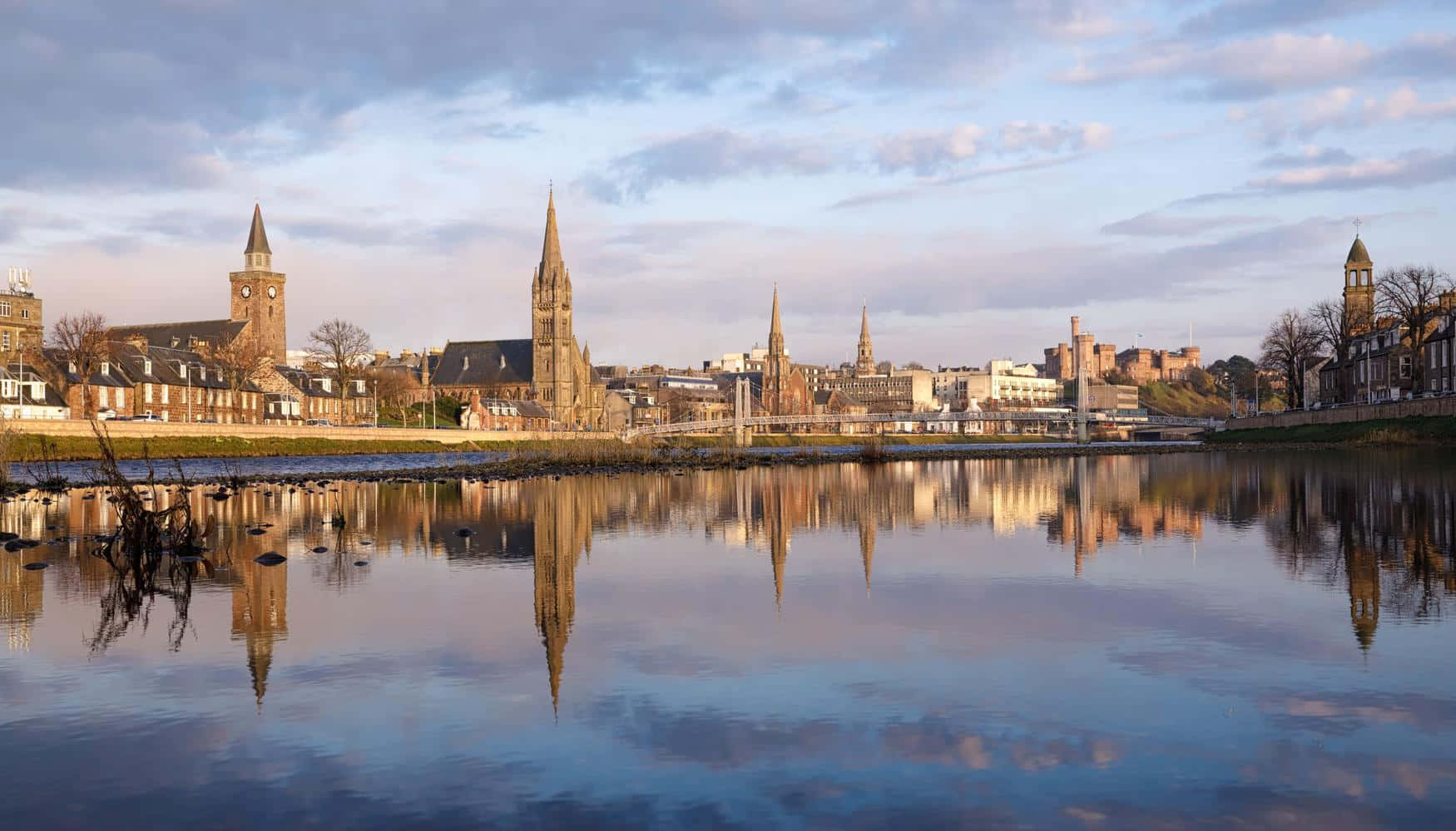 Inverness United Kingdom 桌布 壁紙 — 一條寧靜的河流映照著歷史悠久的石造尖塔和建築，天空柔和暮色，雲朵散佈。