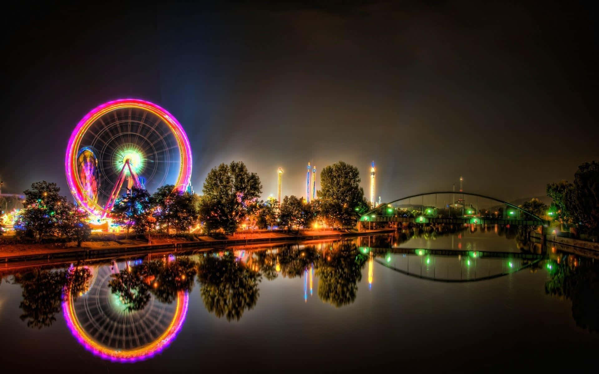  壁紙 — 遊樂園中迷人的夜景，明亮的摩天輪在平靜的水面上倒映，旁邊是一座發光的橋樑。
