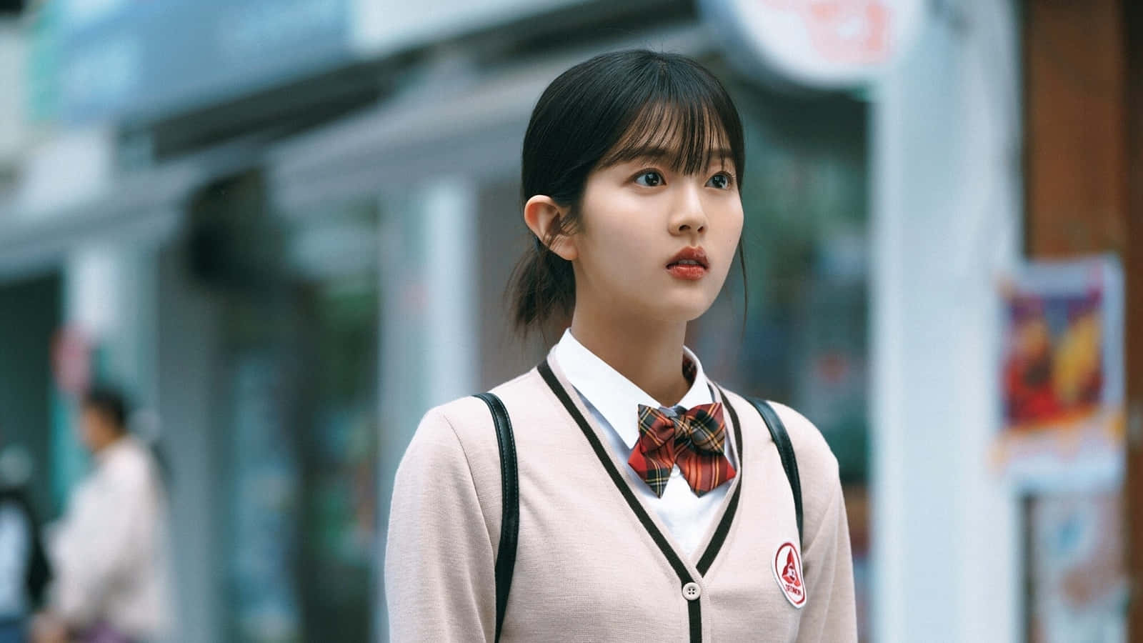  壁紙 — 一個穿著校服、繫著格子蝴蝶結的女孩站在城市街道上，若有所思地望向一旁。