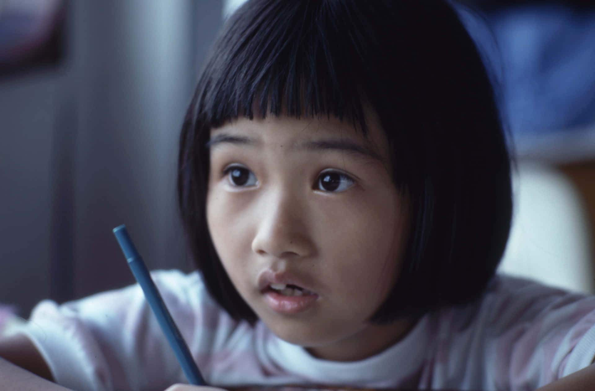  壁紙 — 一位留著深色鮑伯頭和瀏海的年輕女孩，在柔和光線的室內環境中，拿著一支藍色筆，睜著大大的好奇眼睛看向鏡頭外。