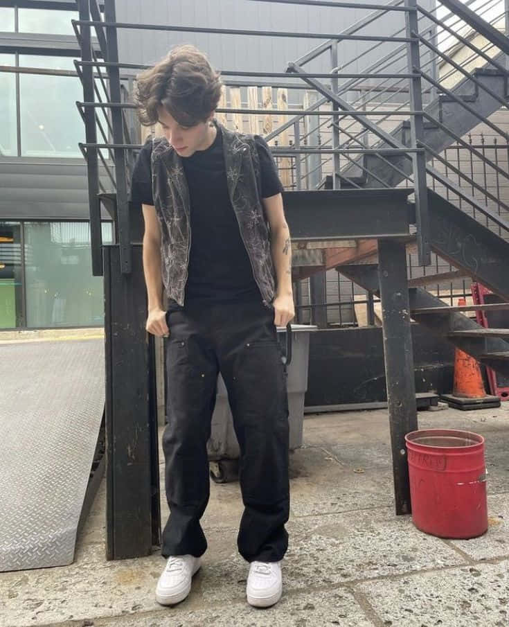  壁紙 — 一名身穿黑色服裝和白色運動鞋的年輕男子，在城市小巷中靠著金屬樓梯，附近有一個紅色水桶。