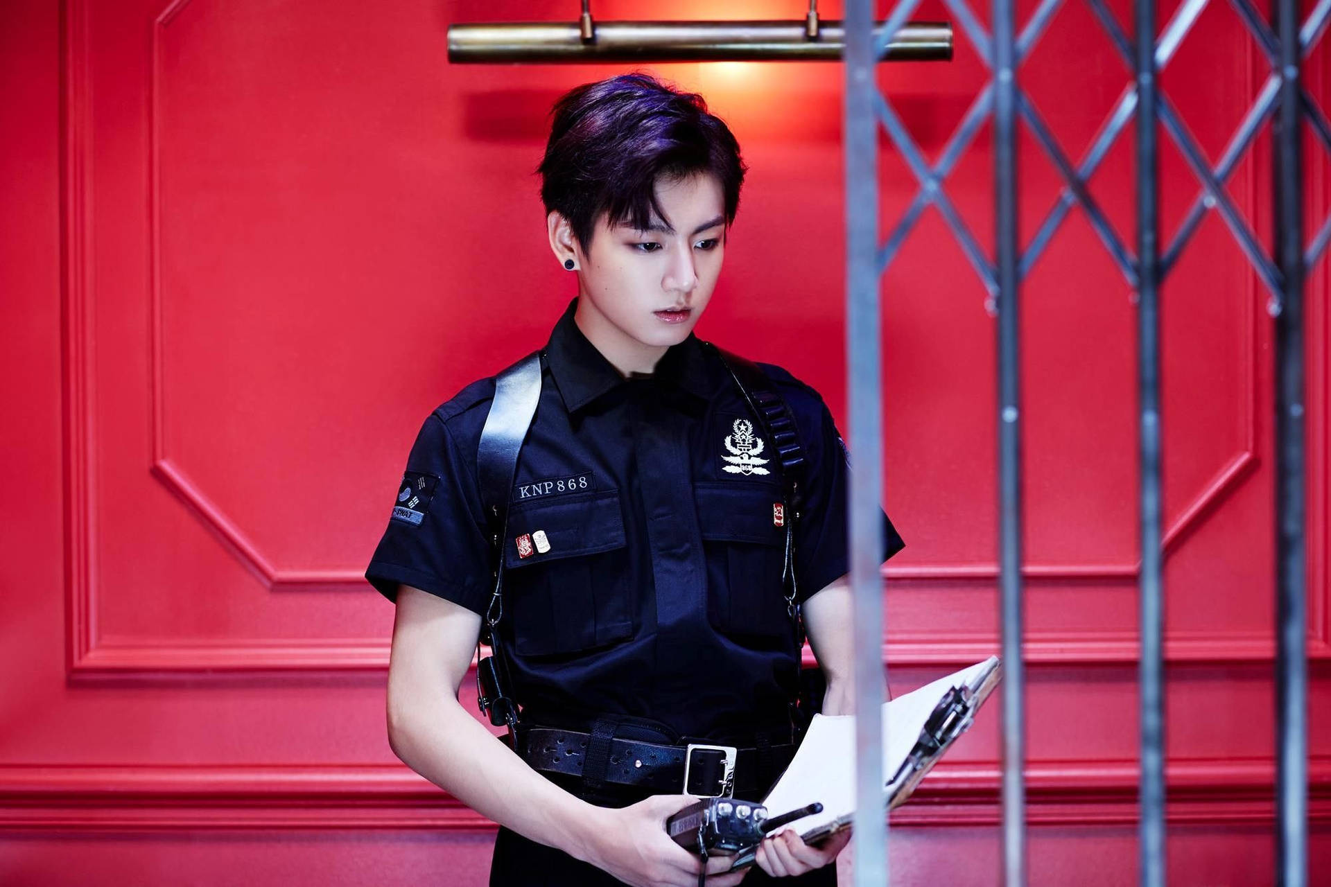 年輕警察在紅色牆前欄杆後手持寫字板。 — Jungkook 壁紙