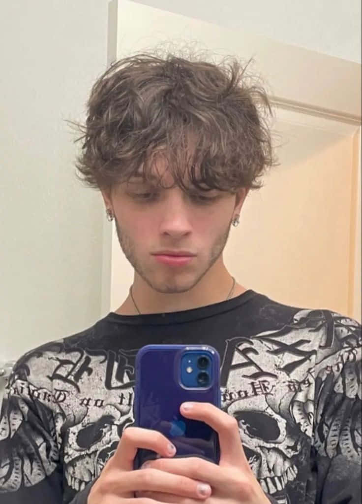  壁紙 — 一個棕色亂髮、穿著骷髏印花襯衫的年輕男子，用藍色手機在鏡子前自拍。