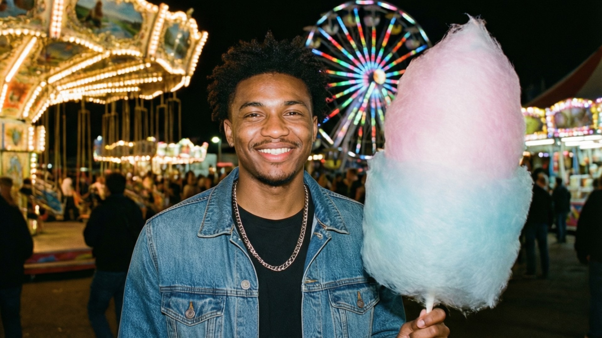 Carnival Night Fun 壁紙 — 一位微笑的年輕男子穿著牛仔外套，在充滿活力的夜間嘉年華中，身後有發光的遊樂設施，手持粉紅色和藍色的棉花糖。
