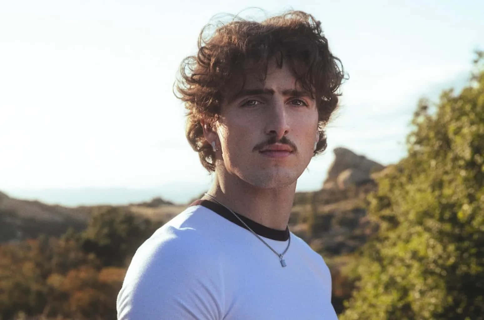  壁紙 — 一位留著棕色捲髮和鬍子的年輕男子，在陽光照耀的山丘背景下平靜地凝視著鏡頭。