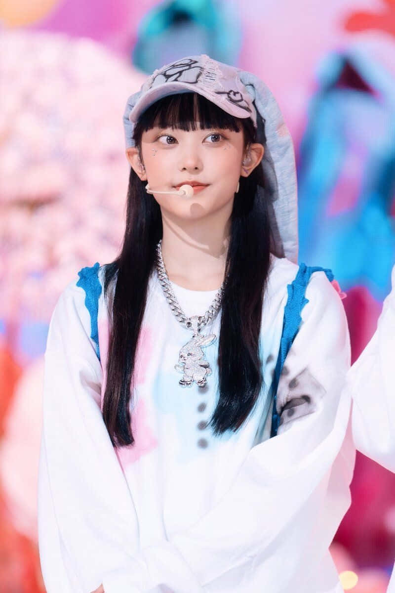  壁紙 — 一位長黑髮的年輕女性戴著圖案帽子和帶有藍色點綴的白色連帽衫，站在燈光明亮的舞台上，背景色彩繽紛且模糊。