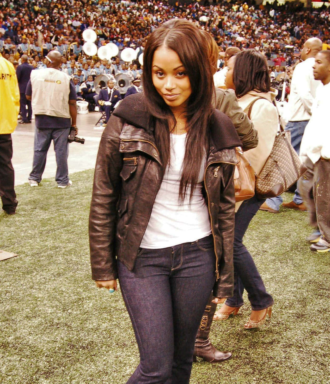 Lauren London 桌布 壁紙 — 一位年輕女性穿著棕色皮夾克和牛仔褲，自信地站在擁擠體育場活動的人造草坪上。