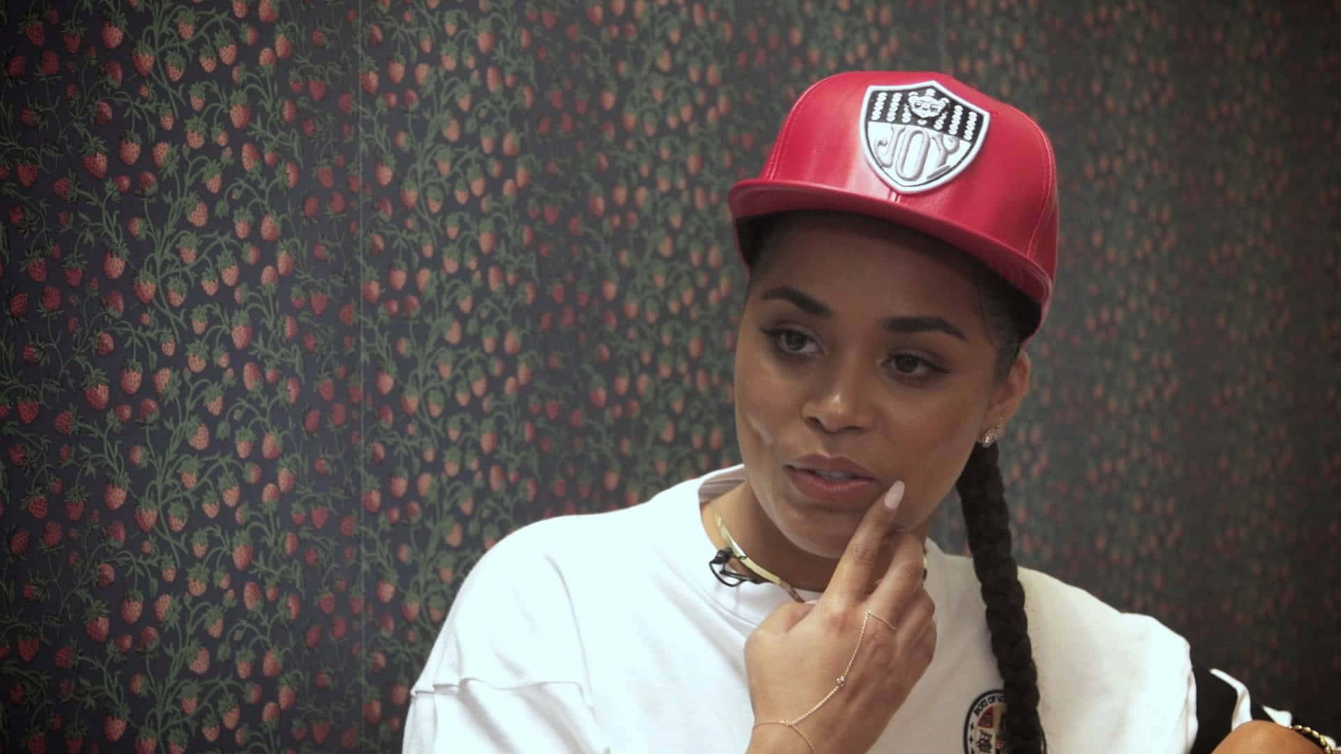 Lauren London 桌布 壁紙 — 一位戴紅帽穿白T恤的年輕女子，手指托著下巴，靠著深色草莓圖案壁紙。