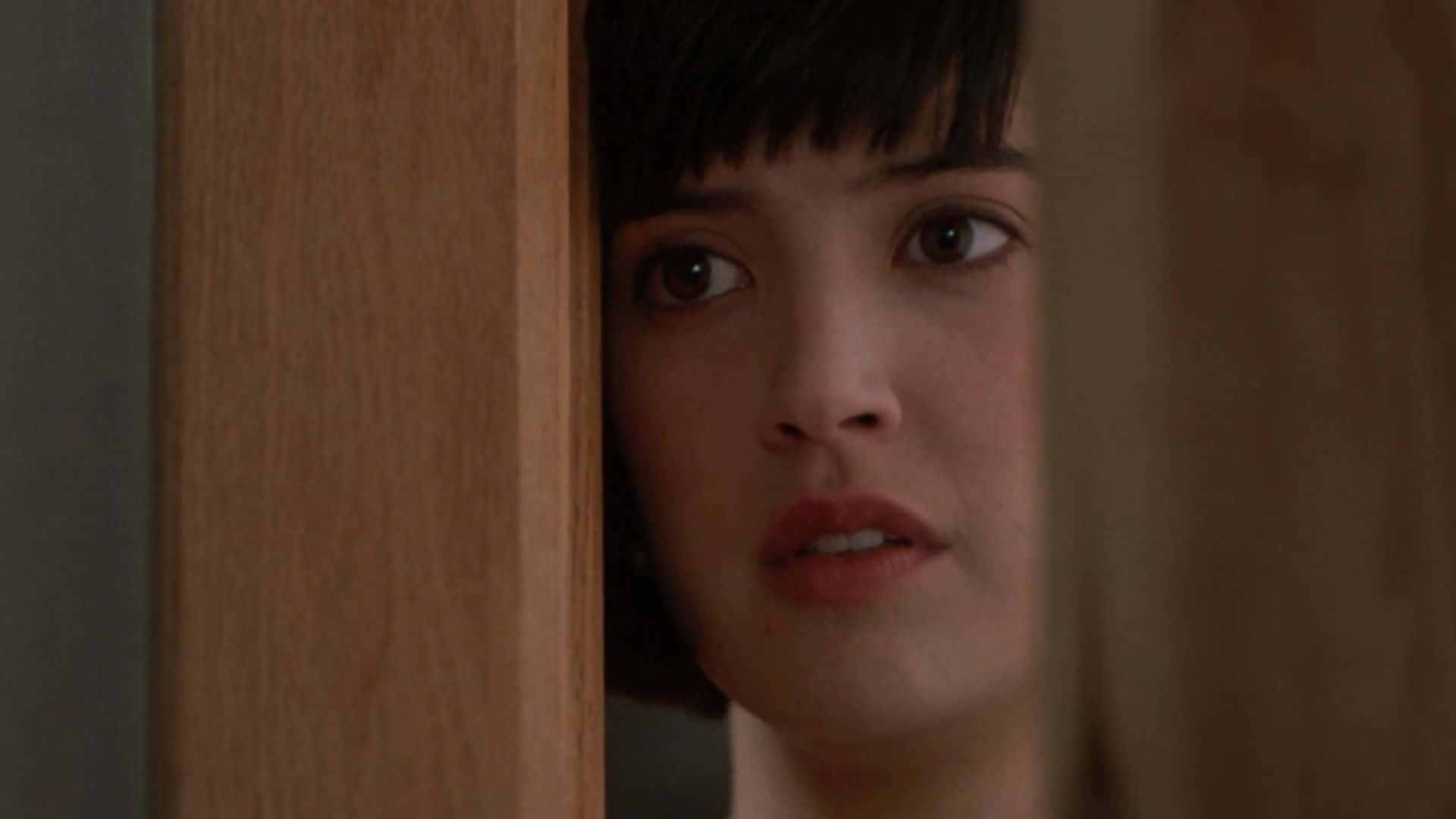 Phoebe Cates 桌布 壁紙 — 一位留著深色鮑伯頭的年輕女子，透過木門框小心翼翼地窺視，睜著一雙充滿好奇的大眼睛。