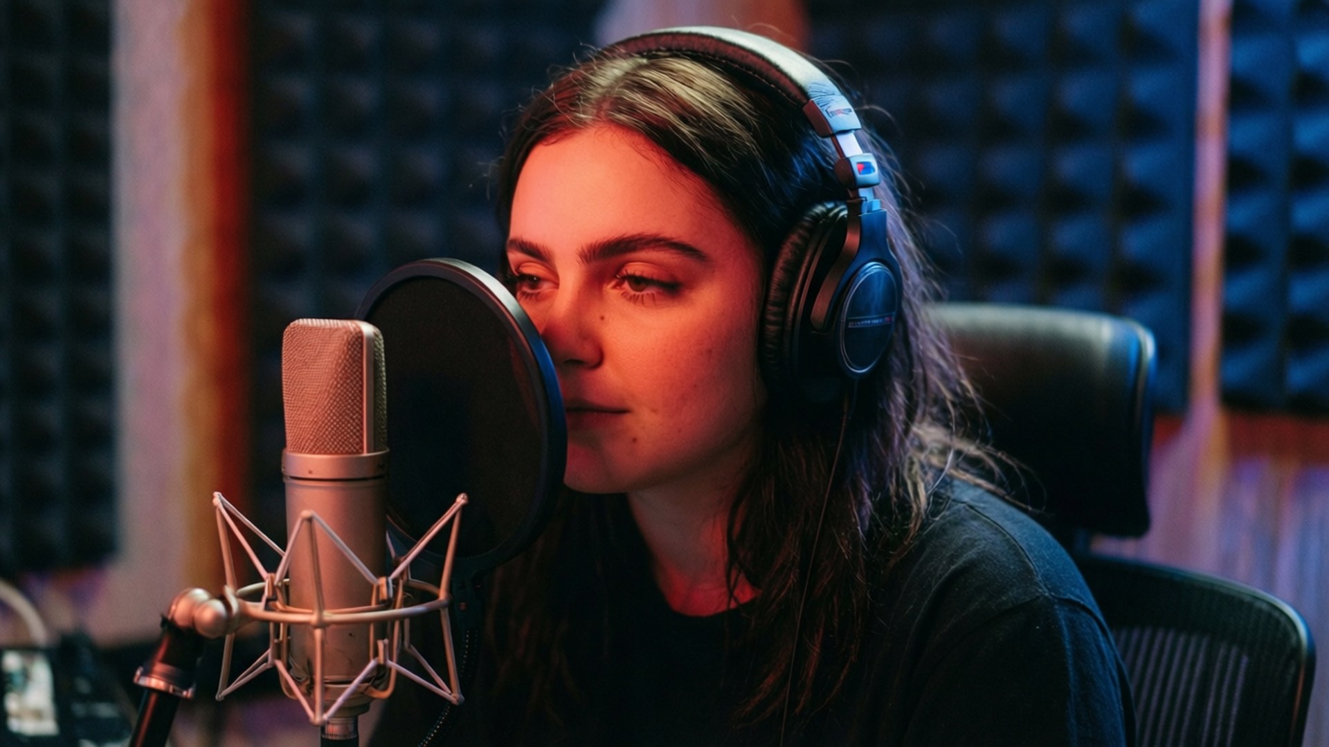 Studio Recording Session 壁紙 — 一位戴著耳機的年輕女性在溫暖的紅色燈光下，對著隔音泡棉牆前的錄音室麥克風唱歌。