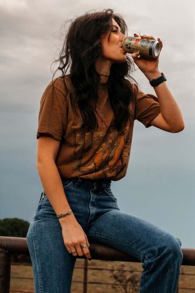 壁紙 — 一位深色頭髮的年輕女性坐在生鏽的欄杆上，在陰天啜飲罐裝飲料。