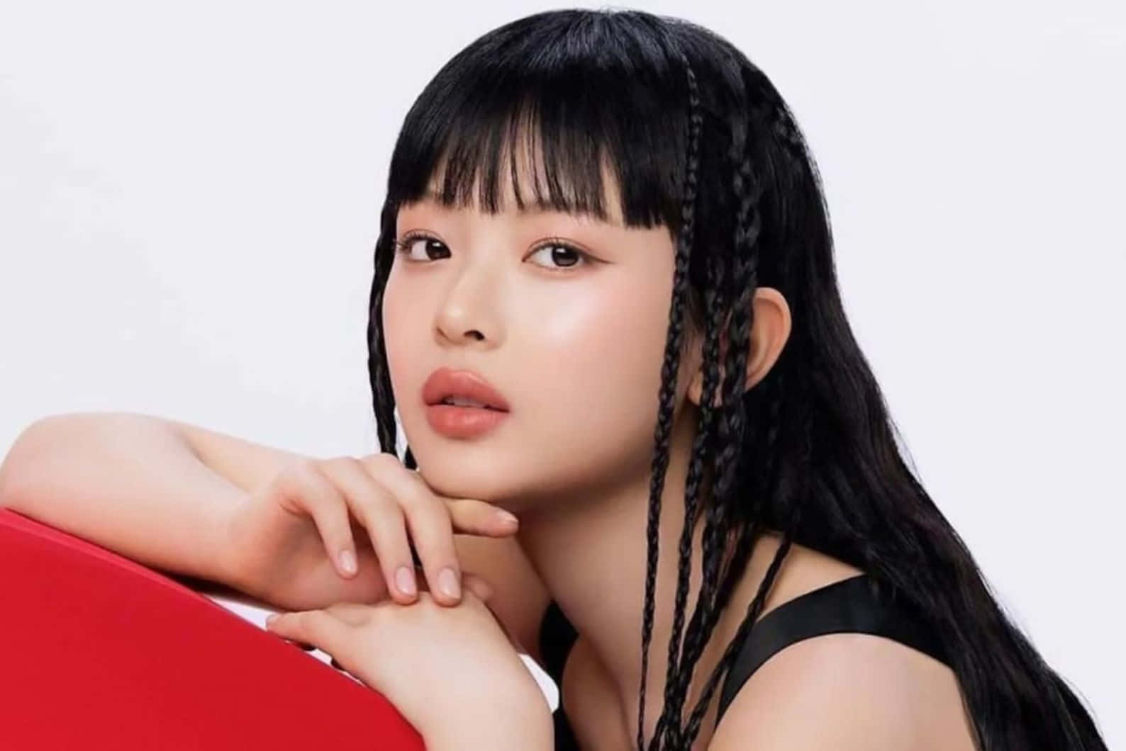  壁紙 — 一位黑色辮子頭髮的年輕女子，在純白色背景上，將下巴擱在雙手之上，靠著大膽的紅色表面。