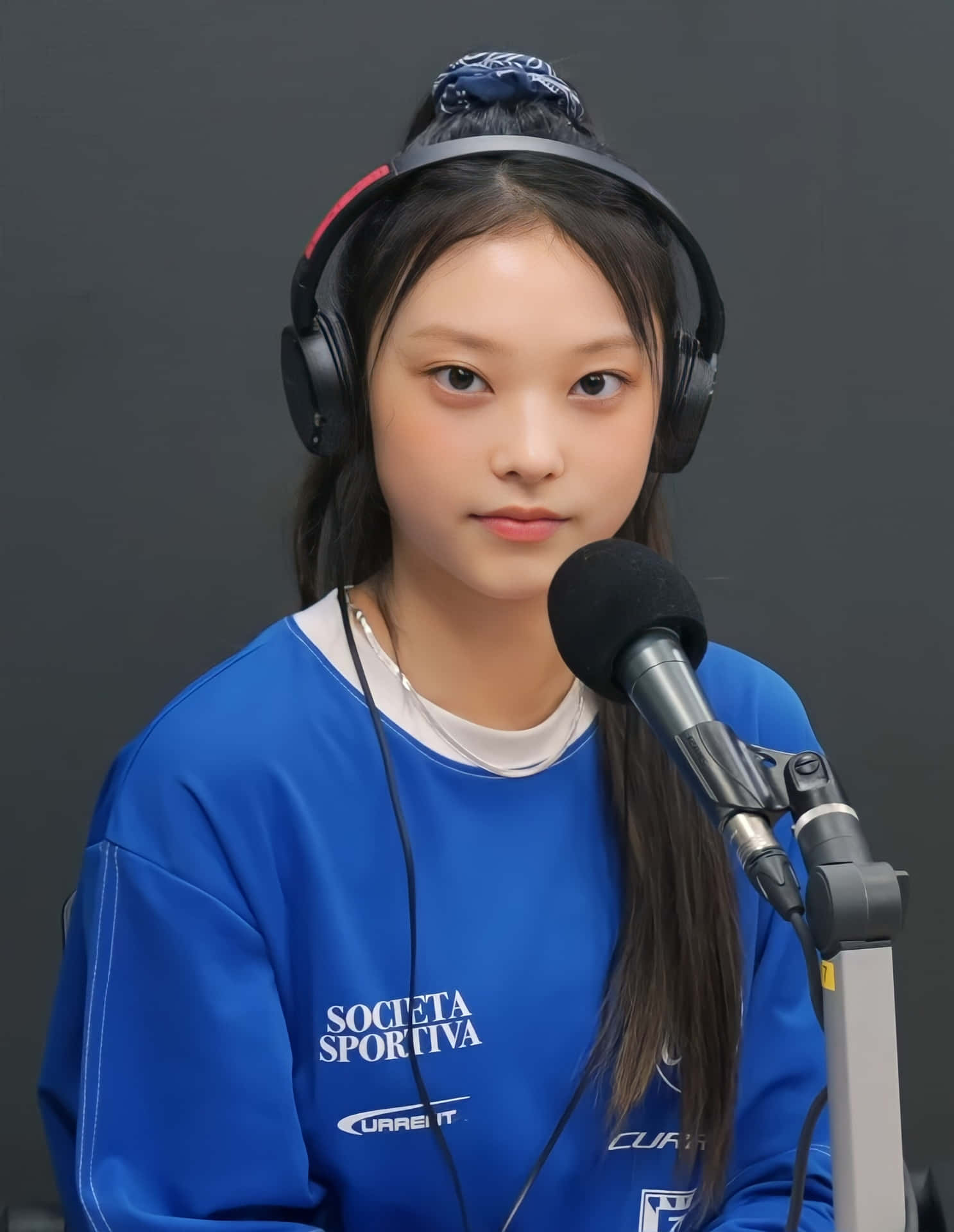  壁紙 — 一位戴著耳機、身穿藍色運動衫的年輕女性坐在麥克風前，背景是深灰色的錄音室。