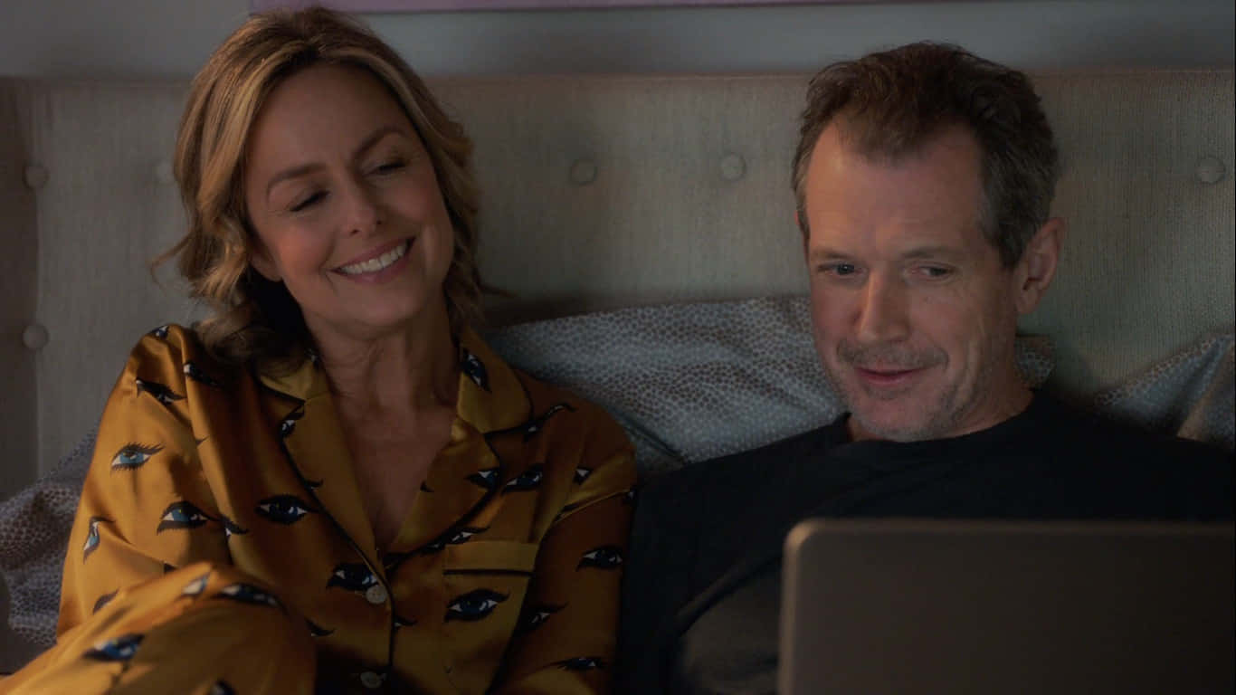 Melora Hardin 桌布 壁紙 — 一對微笑的情侶穿著睡衣在床上放鬆，一起在舒適的臥室裡看筆記型電腦上的東西。