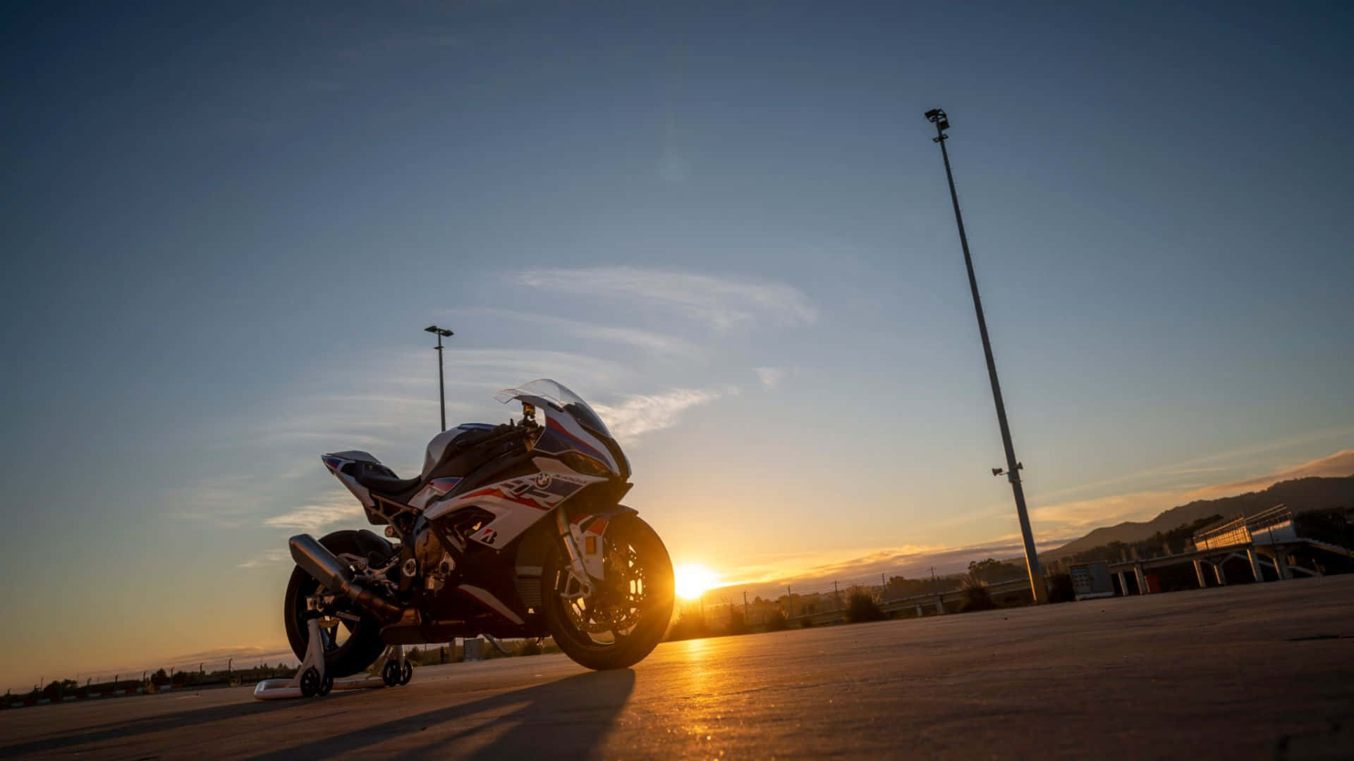  壁紙 — 一輛運動機車停在鋪砌的賽道上，夕陽在它背後落下，投下長長的影子與溫暖的金色光芒。