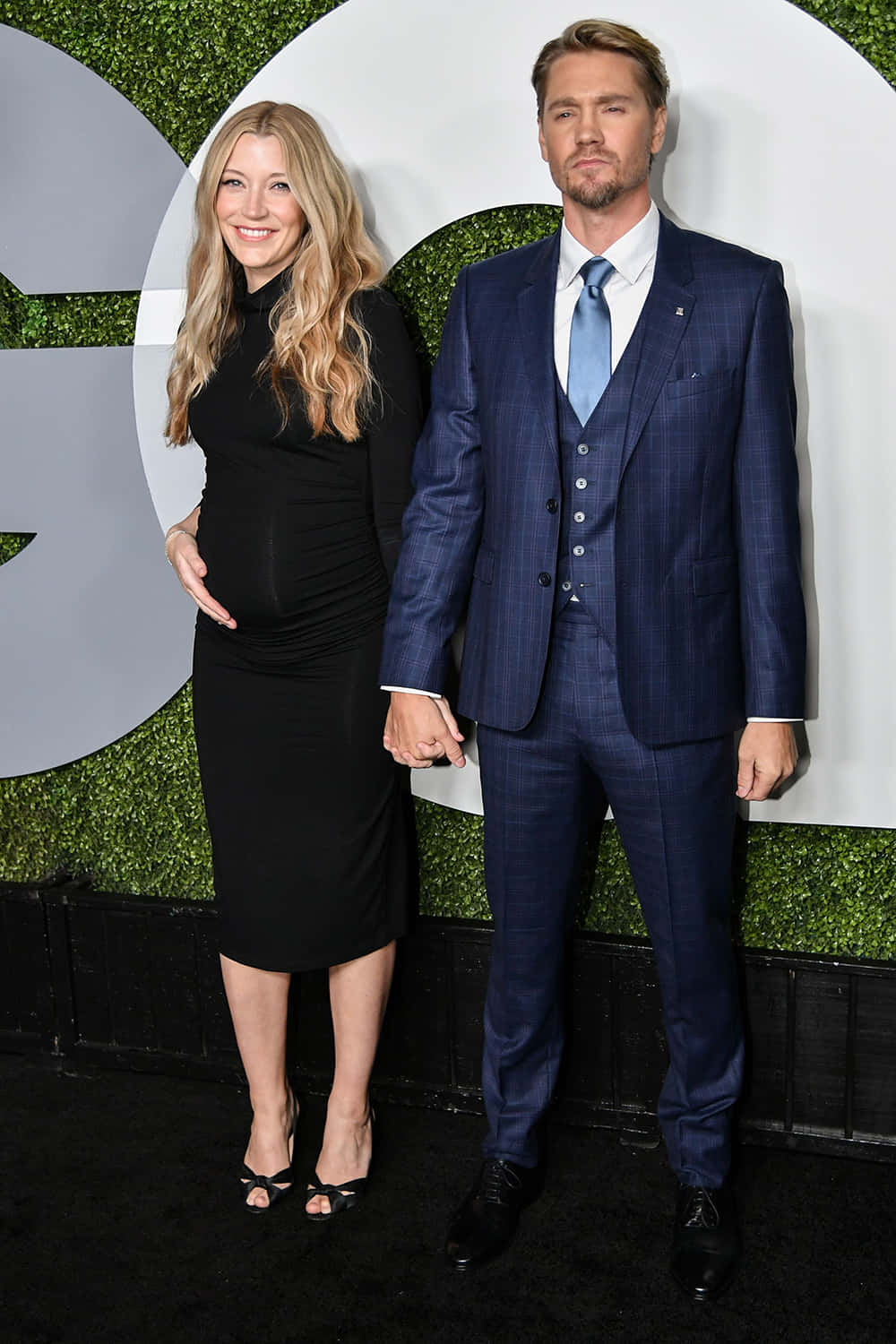 Sarah Roemer 桌布 壁紙 — 一位身穿黑色連衣裙的孕婦，在綠色牆壁活動中，與身穿藍色西裝的伴侶牽手微笑。