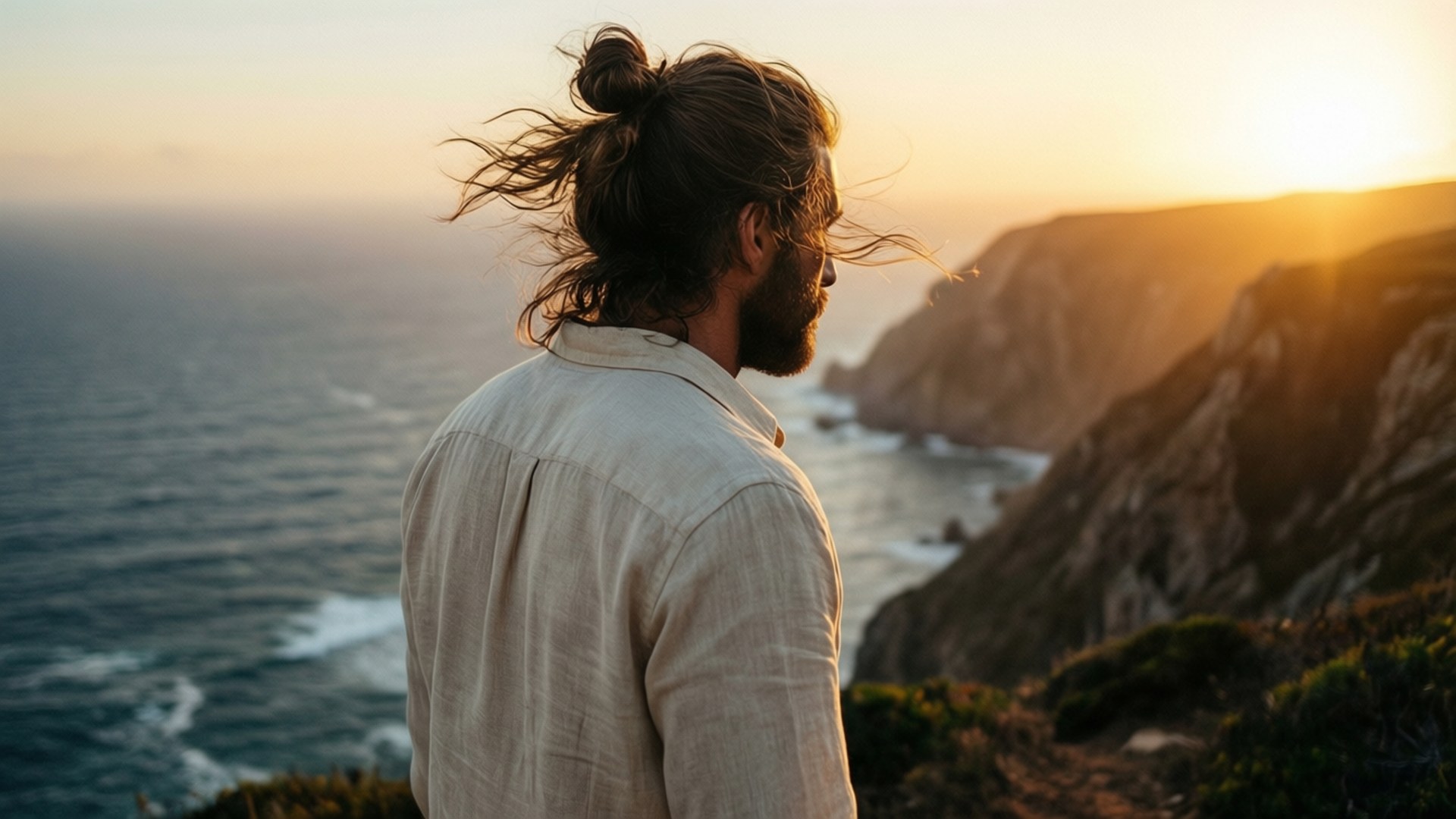 Ocean Sunset 壁紙 — 一個紮髻留鬚的男人站在懸崖上，在日落時分俯瞰海洋，風吹動他的頭髮。