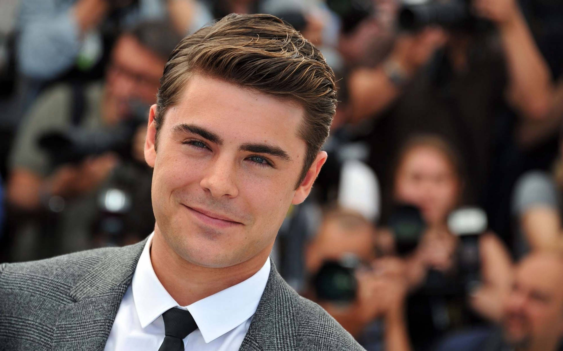Zac 桌布 壁紙 — Zac Efron 穿著灰色西裝，在擁擠的紅毯活動中自信地微笑，身後閃光燈閃爍。