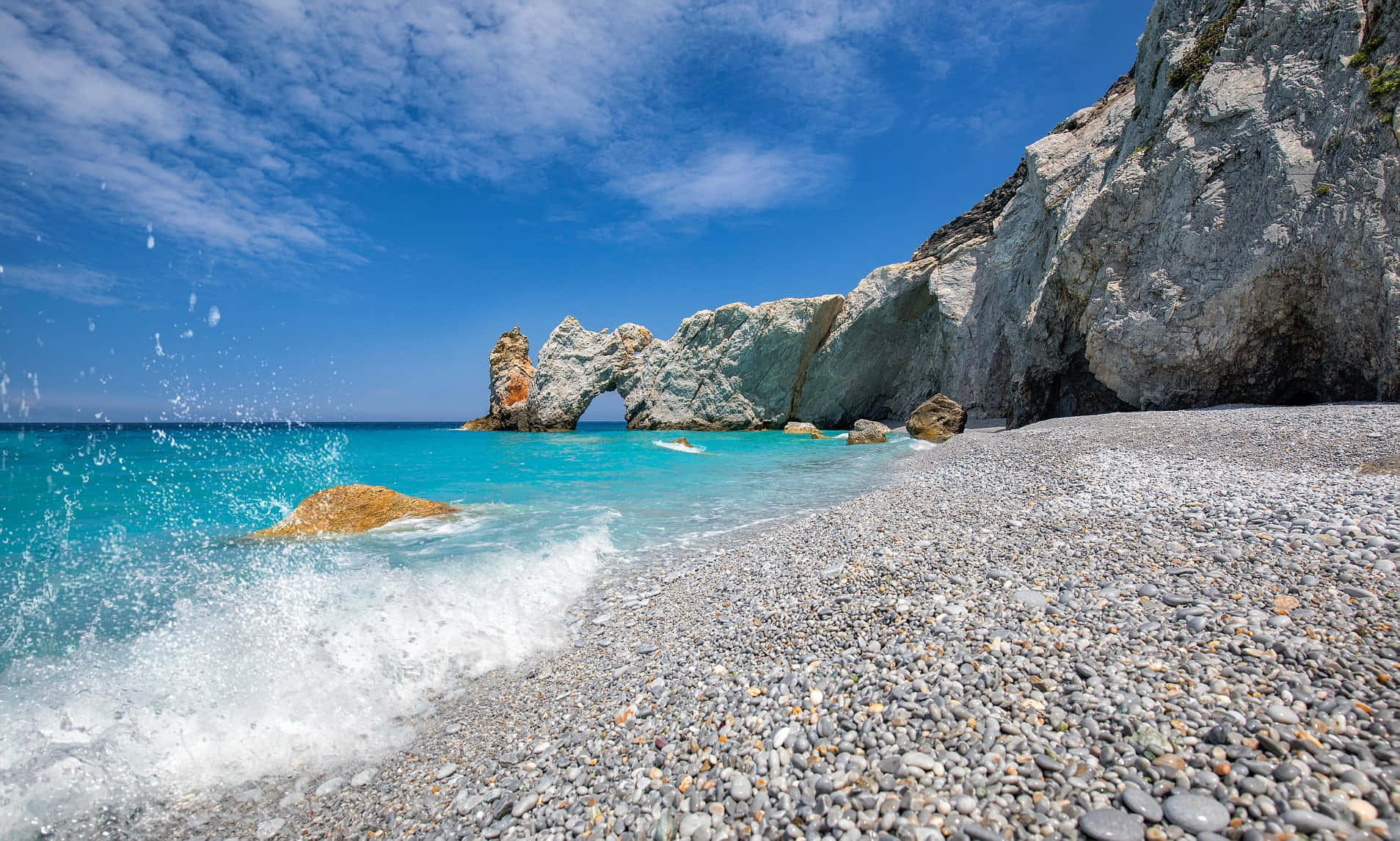 Greek Beach 桌布 壁紙 — 在天然岩石拱門附近，明亮的藍天白雲下，碧綠色的波浪拍打著鵝卵石海灘。