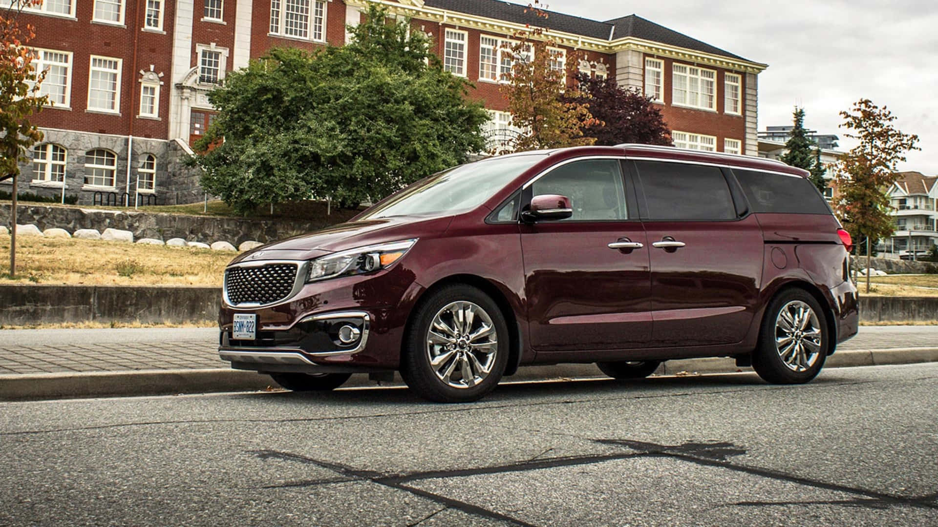 Kia Sedona 桌布 壁紙 — 一輛棗紅色小型貨車停在一條街上，背景有磚砌建築和秋天的樹木。