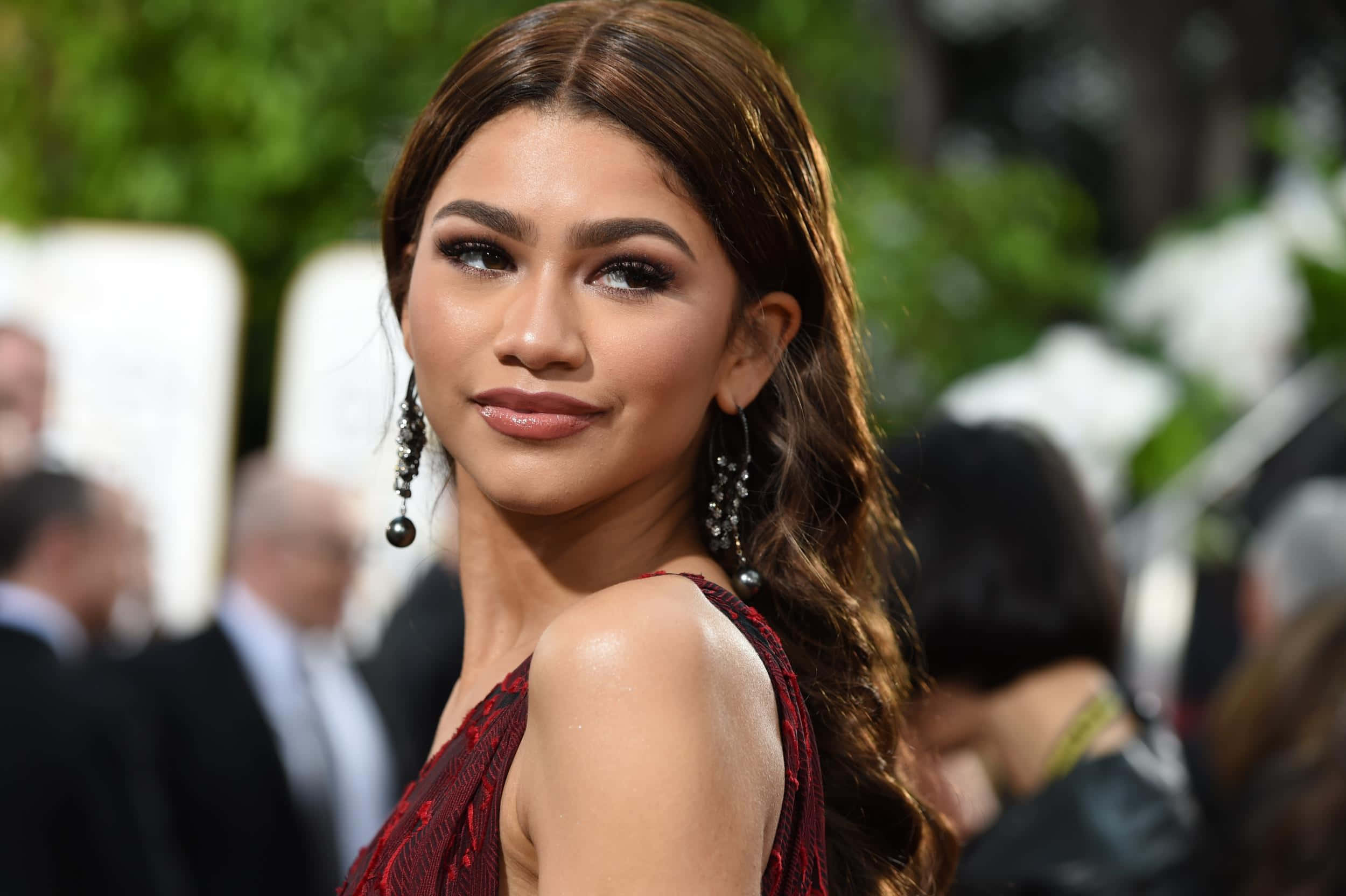 Celebrity 桌布 壁紙 — Zendaya 在戶外活動中優雅地擺姿勢，身穿紅色連衣裙，戴著垂墜耳環，背景是模糊的綠植和賓客。