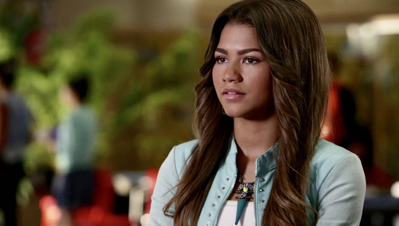 Zapped 桌布 壁紙 — Zendaya 在柔和光線的室內環境中若有所思地凝視，身後是模糊的綠色植物和溫暖色調。