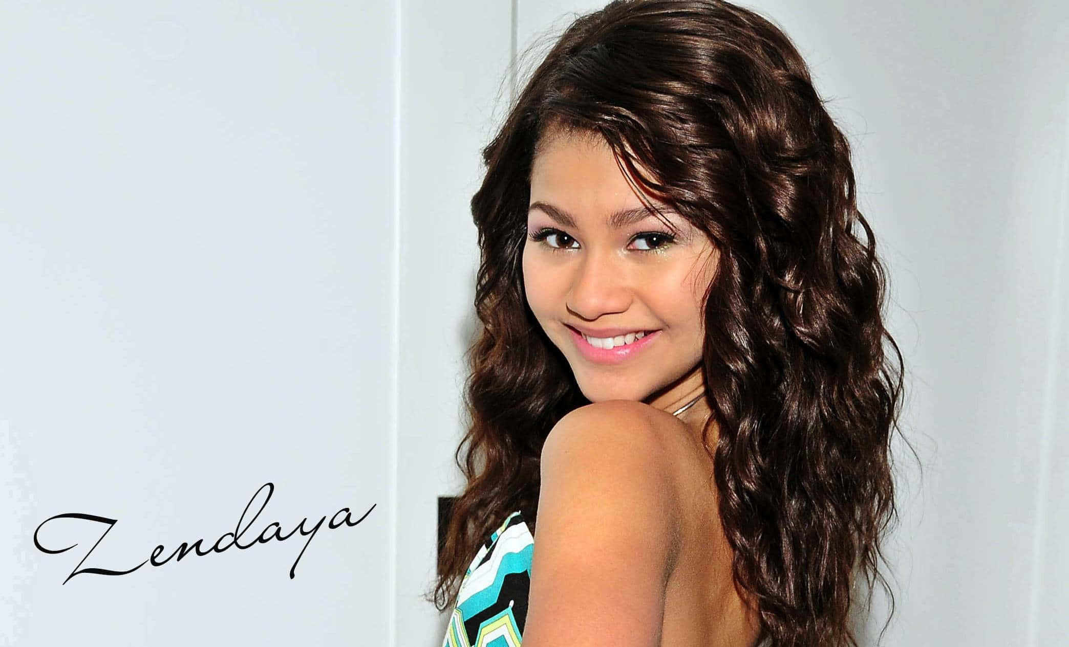 贊達亞 桌布 壁紙 — Zendaya 在純白色背景前，留著長長的波浪形棕色頭髮，露出溫暖的笑容。