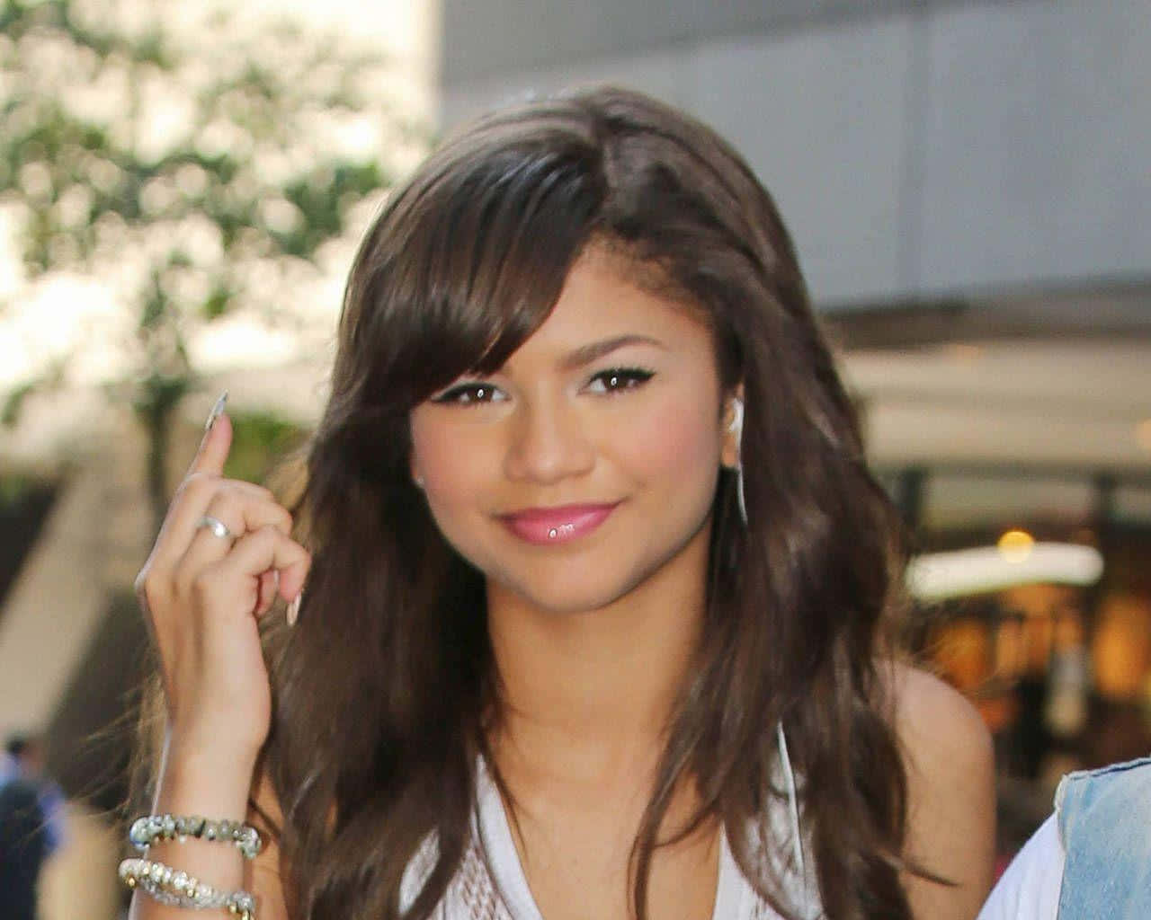 Zendaya 桌布 壁紙 — Zendaya 微笑著面對鏡頭，長長的棕色頭髮，手指以俏皮的姿勢舉起。