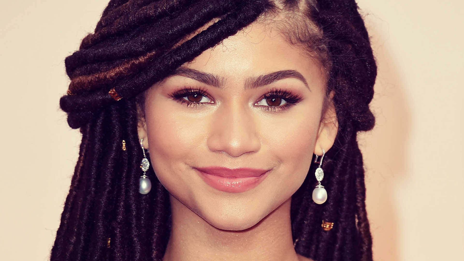 Zendaya 桌布 壁紙 — Zendaya 溫暖微笑，長長的黑辮子飾有金色珠子和珍珠耳環，背景是柔和的米色。