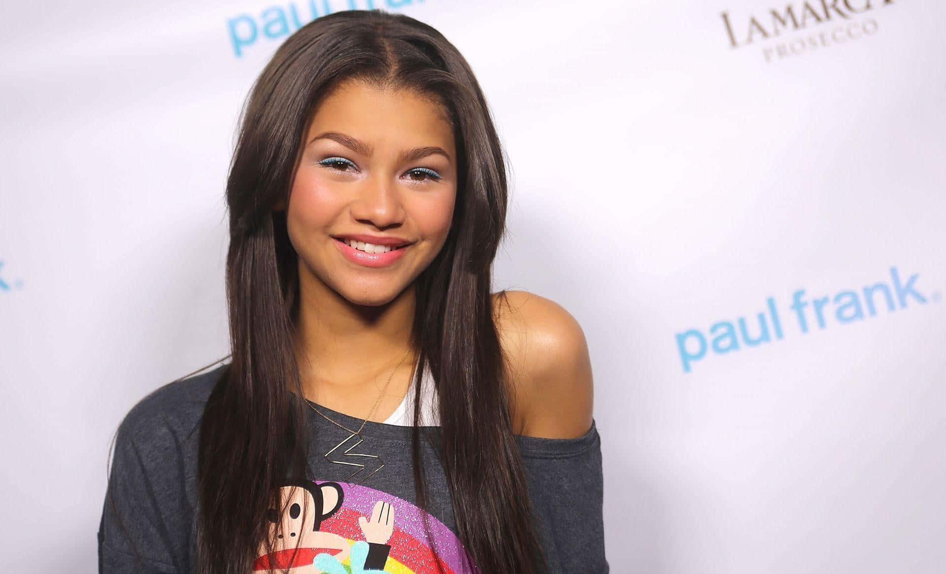 Zendaya 桌布 壁紙 — Zendaya 穿著灰色露肩T恤，在品牌活動背景前溫暖微笑。