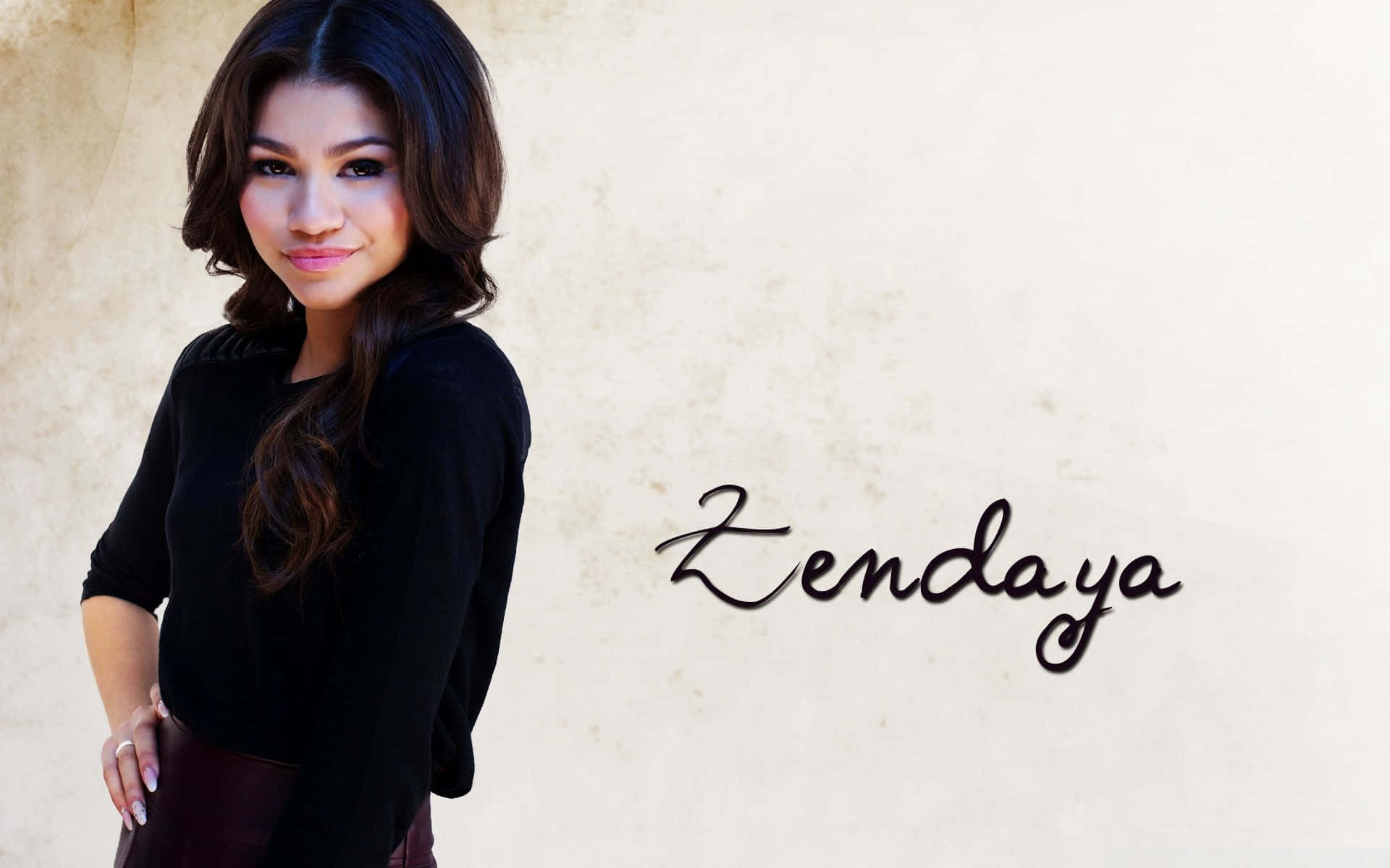 Celebrity 桌布 壁紙 — Zendaya 自信地穿著黑色上衣，站在有紋理的米色牆前，旁邊寫著她的名字。