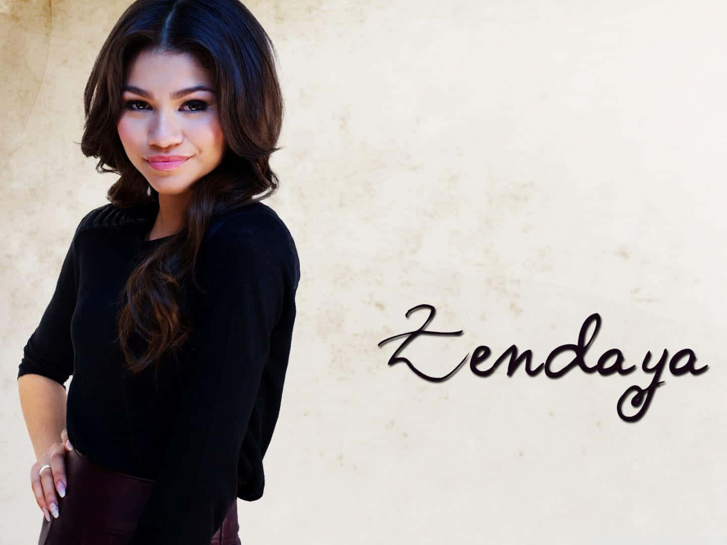 Zendaya 桌布 壁紙 — Zendaya 自信地穿著黑色上衣，靠在有紋理的米色牆上，旁邊寫著她的名字。