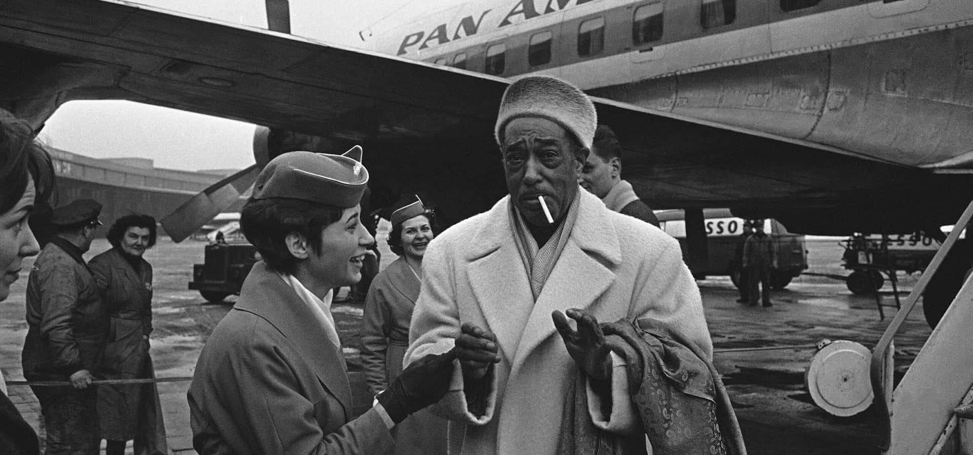  壁紙 — 一張復古黑白照片顯示，一位微笑的泛美航空空服員在機場停機坪上，向一位穿著大衣、戴著帽子、抽著香菸的男子打招呼。